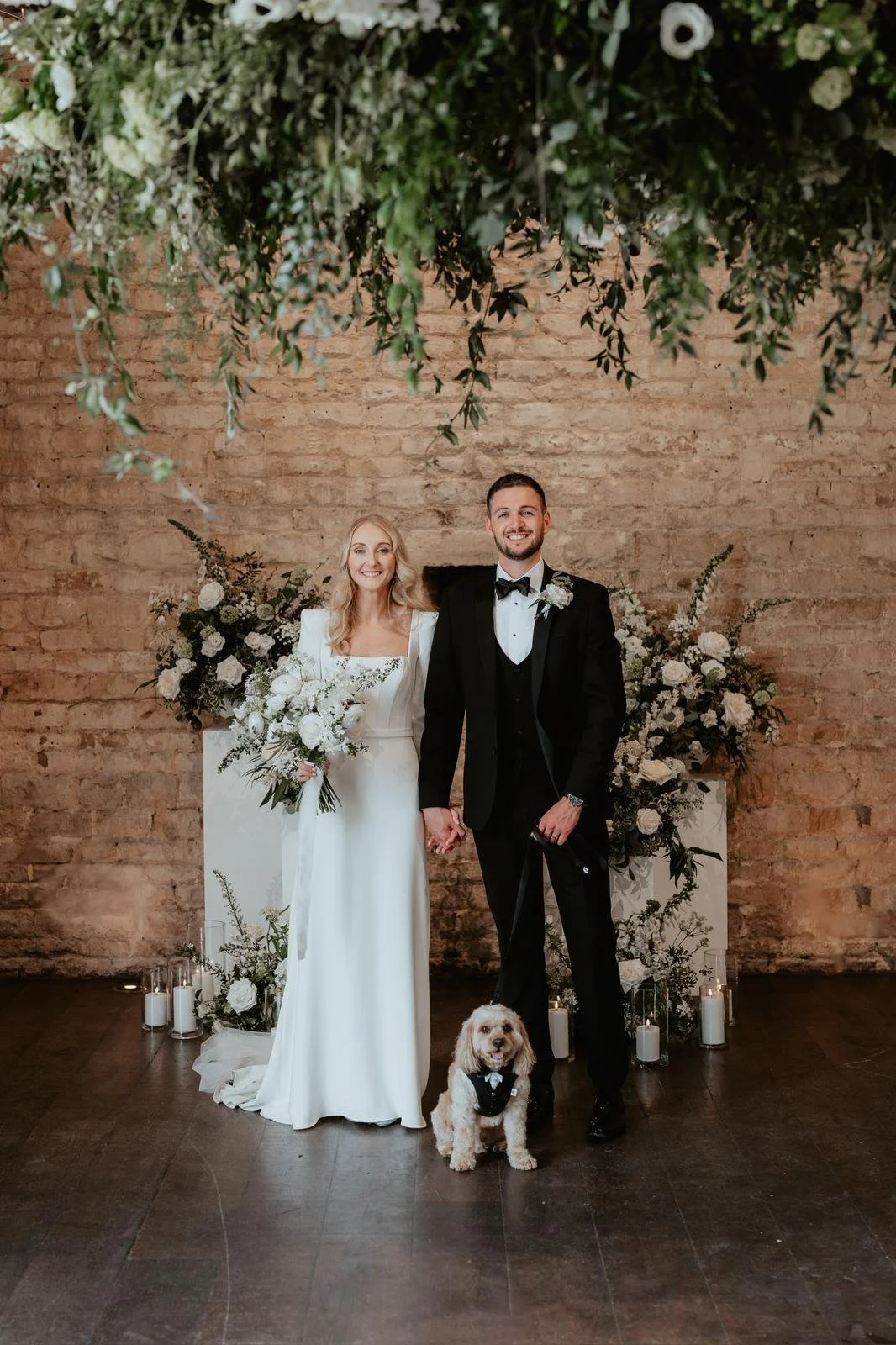 A bride and groom holding hands, smiling at the camera, standing with a dog dressed in a tuxedo, in front of a floral backdrop with candles on the floor, brick wall background, during their wedding celebration.