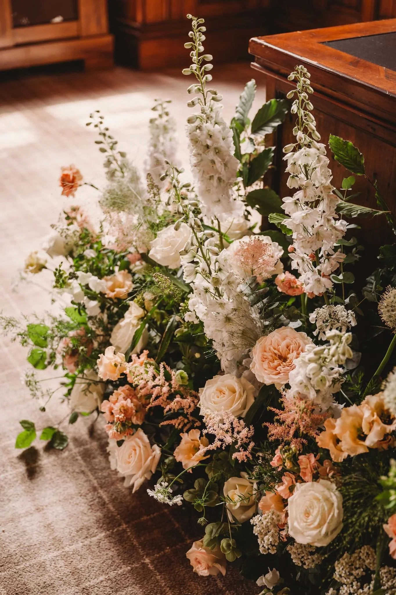 A large, colorful floral arrangement with white, peach, and pink flowers, including roses, snapdragons, and other blooms, placed on a carpeted floor near a wooden piece of furniture.
