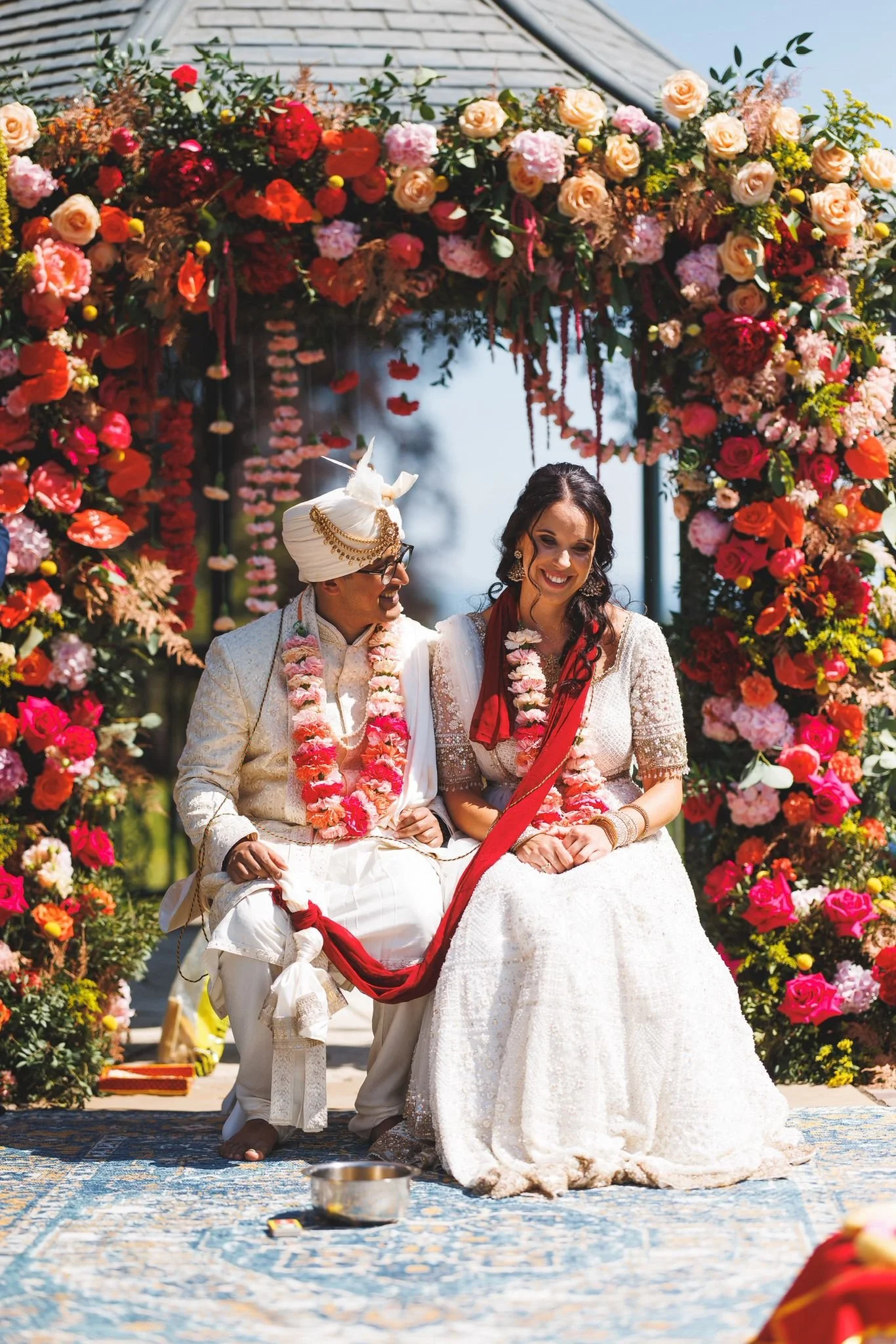 A newlywed couple of different cultural backgrounds, dressed in traditional wedding attire, sitting beneath a vibrant floral arch during an outdoor wedding ceremony, smiling and holding hands.