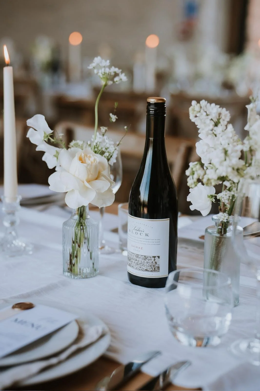 A table set with white flowers in glass vases, a bottle of red wine, a lit candle, and neatly arranged plates and glassware, suggesting an elegant dining setting.