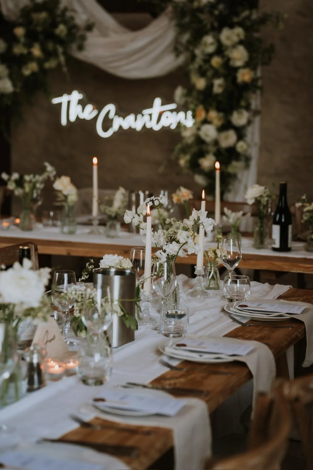 Wedding reception table decorated with white flowers, candles, and glassware, with a neon sign that reads 'The Craytons' in the background.