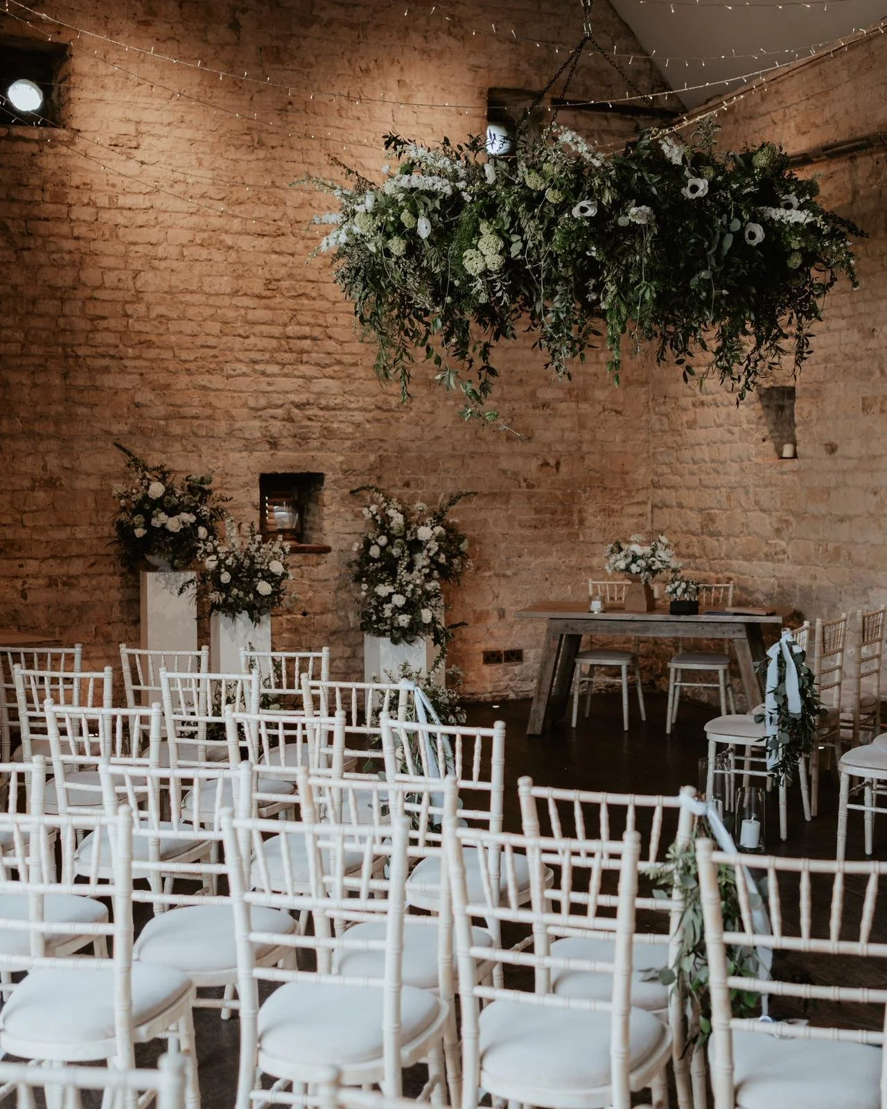 Wedding ceremony setup with white chairs, floral arrangements, and a large hanging floral chandelier in a rustic brick venue.