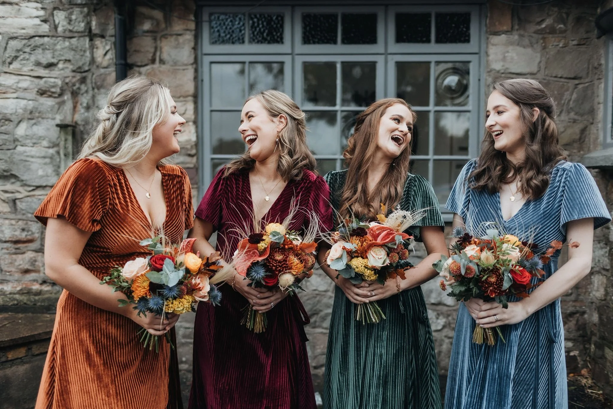 Four women in colorful velvet dresses holding bouquets of flowers, laughing and talking outside against a stone wall.
