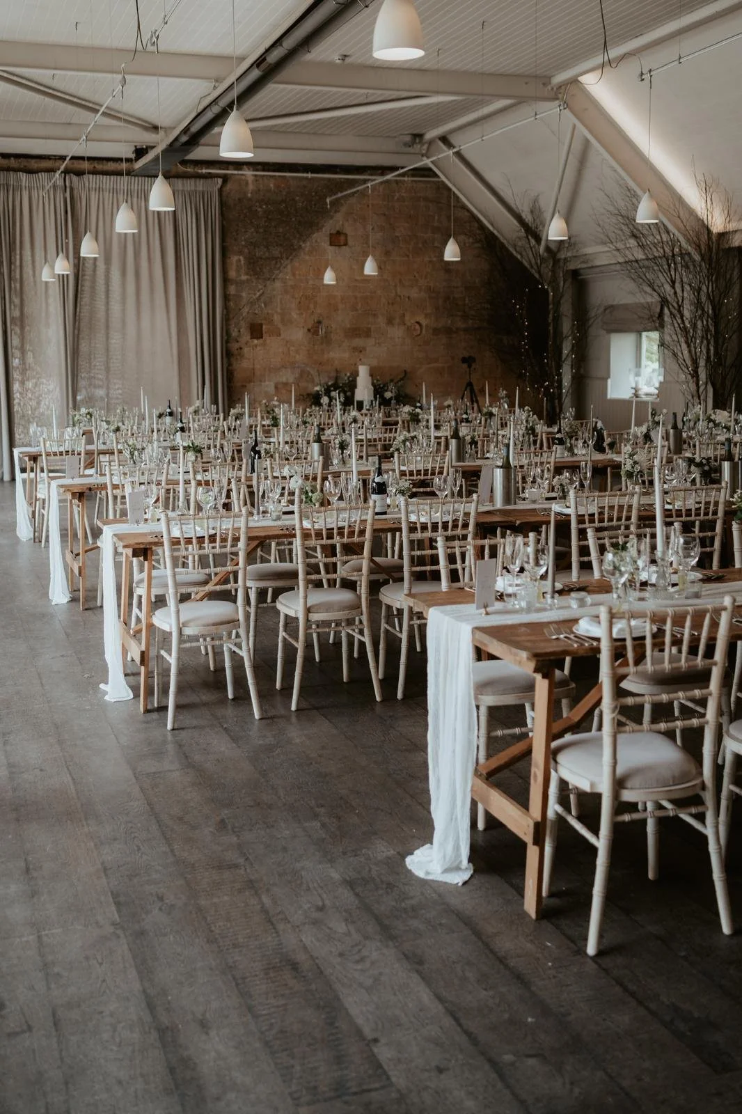 Elegant wedding reception setup in a rustic venue, with long wooden tables, white chairs, and table settings including glassware and floral centerpieces, under hanging pendant lights.