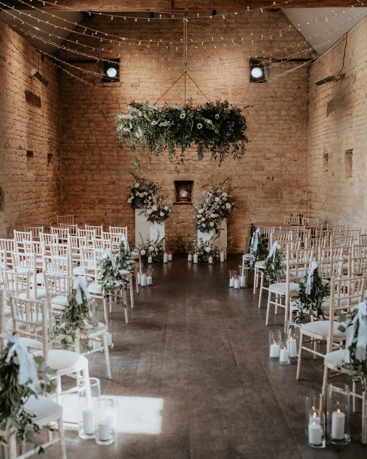 Indoor wedding or event ceremony setup with white chairs decorated with greenery and white ribbons, a floral arrangement hanging from the ceiling, and candles along the aisle, in a rustic brick-walled space.