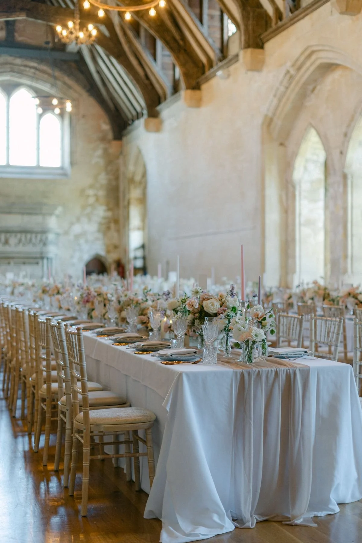 Elegant banquet table decorated with pink and white flowers, candles, and fine china in a historic stone building with large windows and wooden beams.