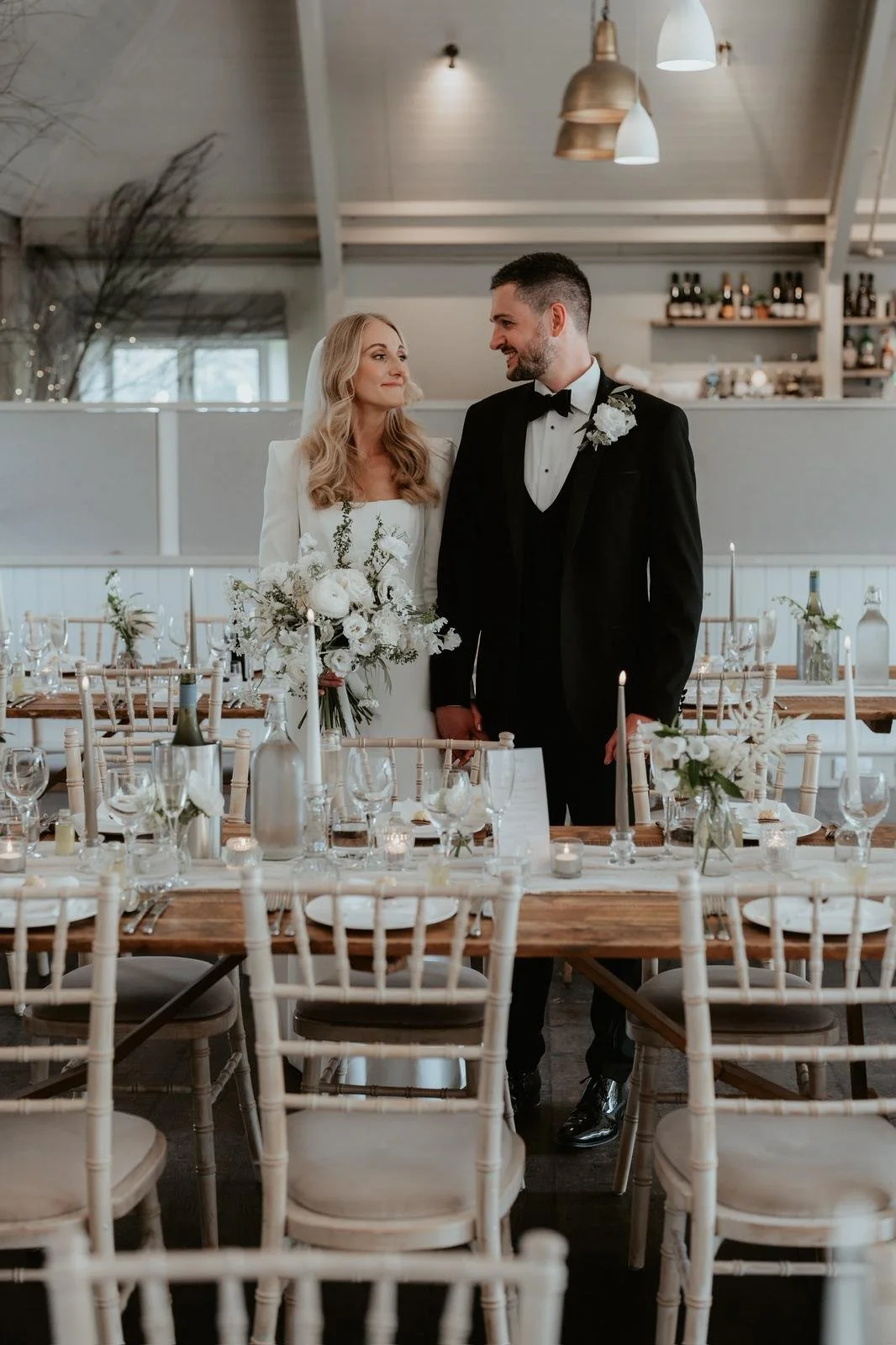 A bride and groom standing together at their wedding reception inside a decorated venue. The bride is holding a bouquet of white flowers and wearing a white wedding dress, while the groom is dressed in a black tuxedo with a white shirt and black bow
