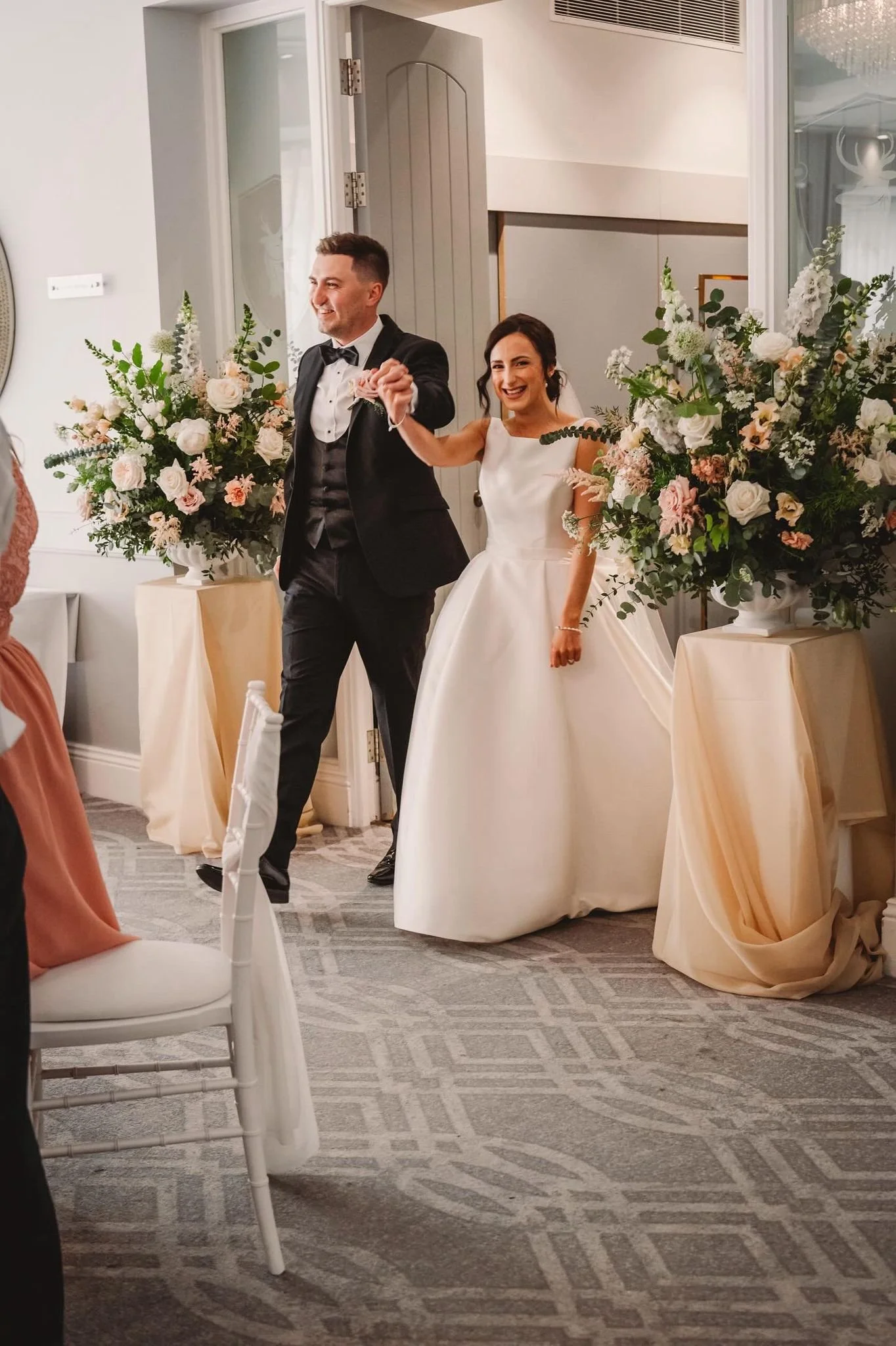 A bride and groom smiling and holding hands as they enter a wedding ceremony, decorated with large floral arrangements. The bride wears a white wedding gown, and the groom is in a tuxedo.