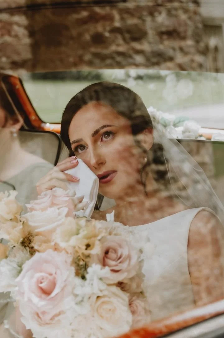 A bride crying, holding a tissue, with a bouquet of pink and white flowers, seen through a reflection in a car window.