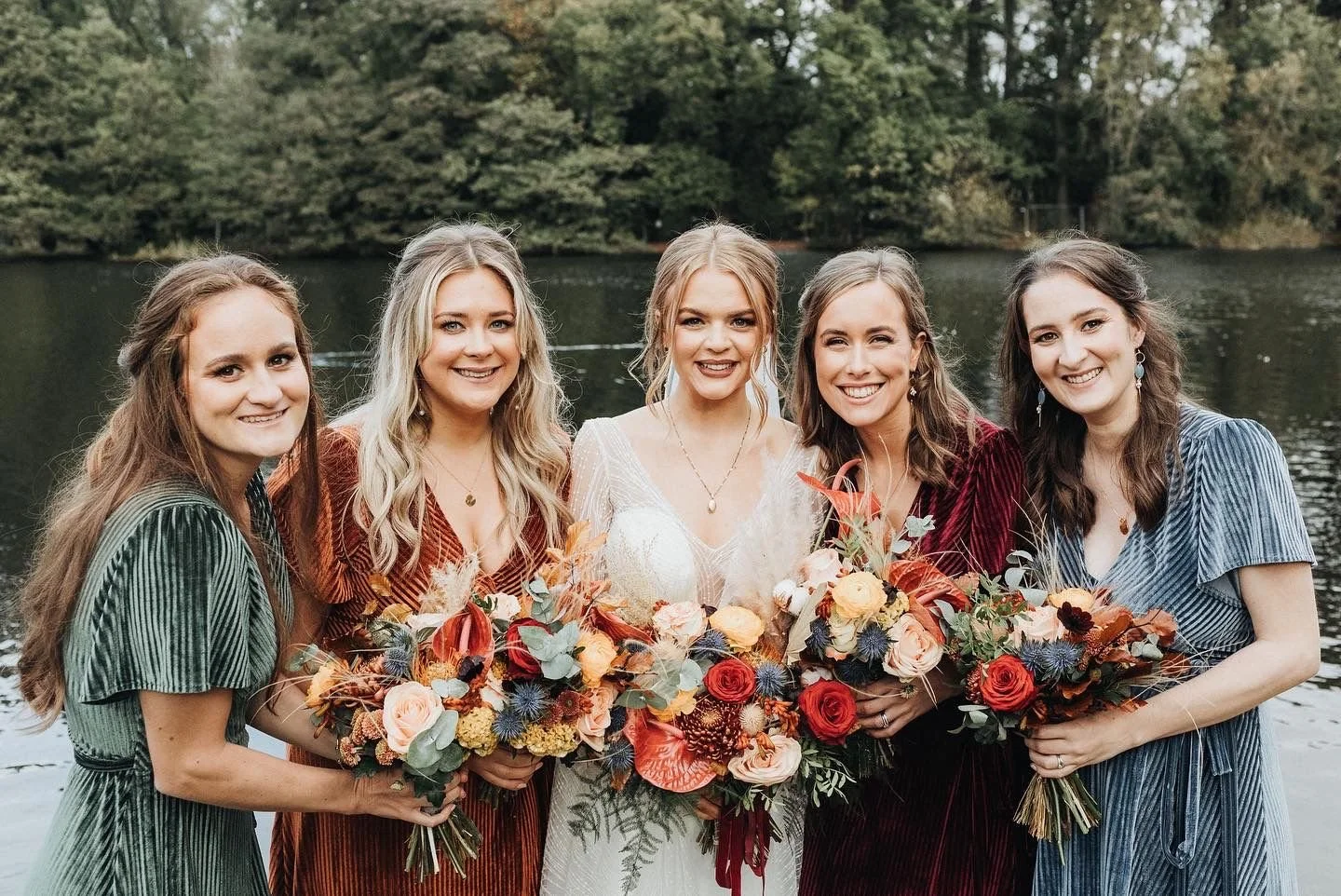 Five women in colorful dresses standing by a lake, holding bouquets of flowers, smiling at the camera during a wedding celebration.
