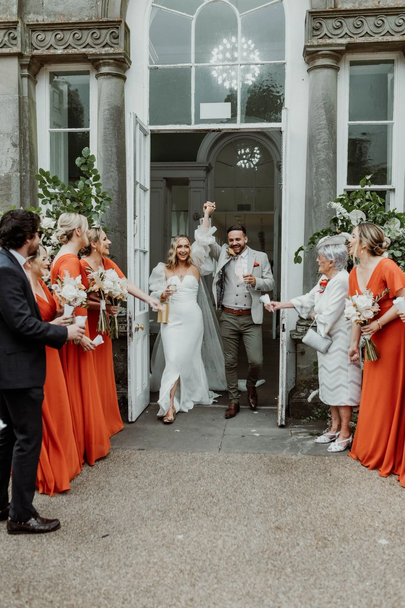 Happy bride and groom exiting a building, celebrating with guests outside, with flowers and greenery in the background.
