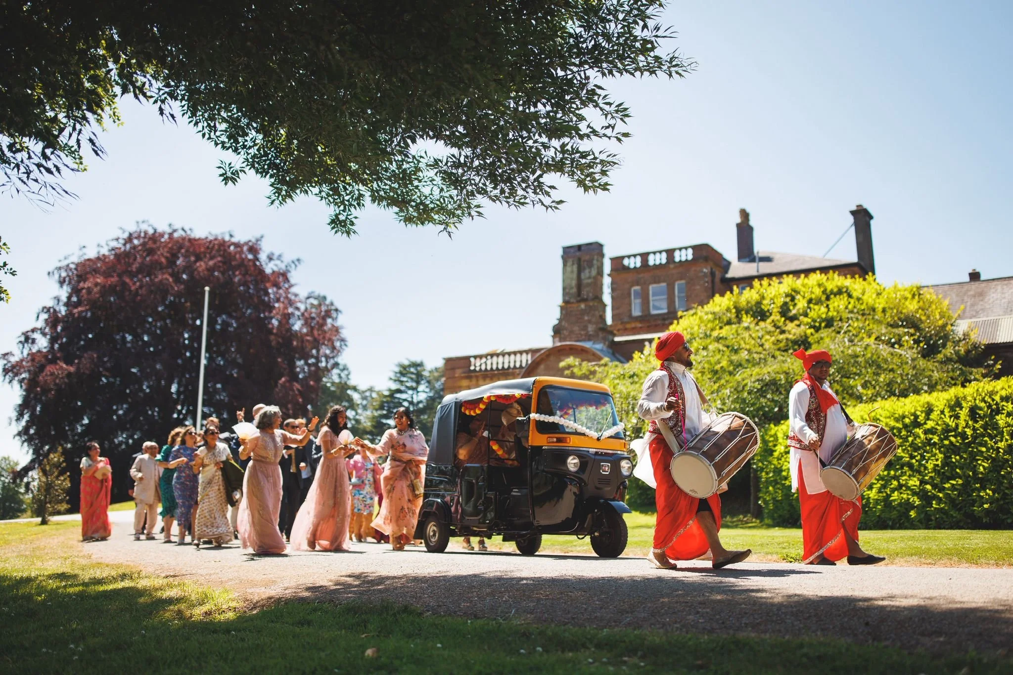 People in colorful traditional Indian attire walking in a parade with musicians playing drums, surrounded by greenery and trees on a bright sunny day.