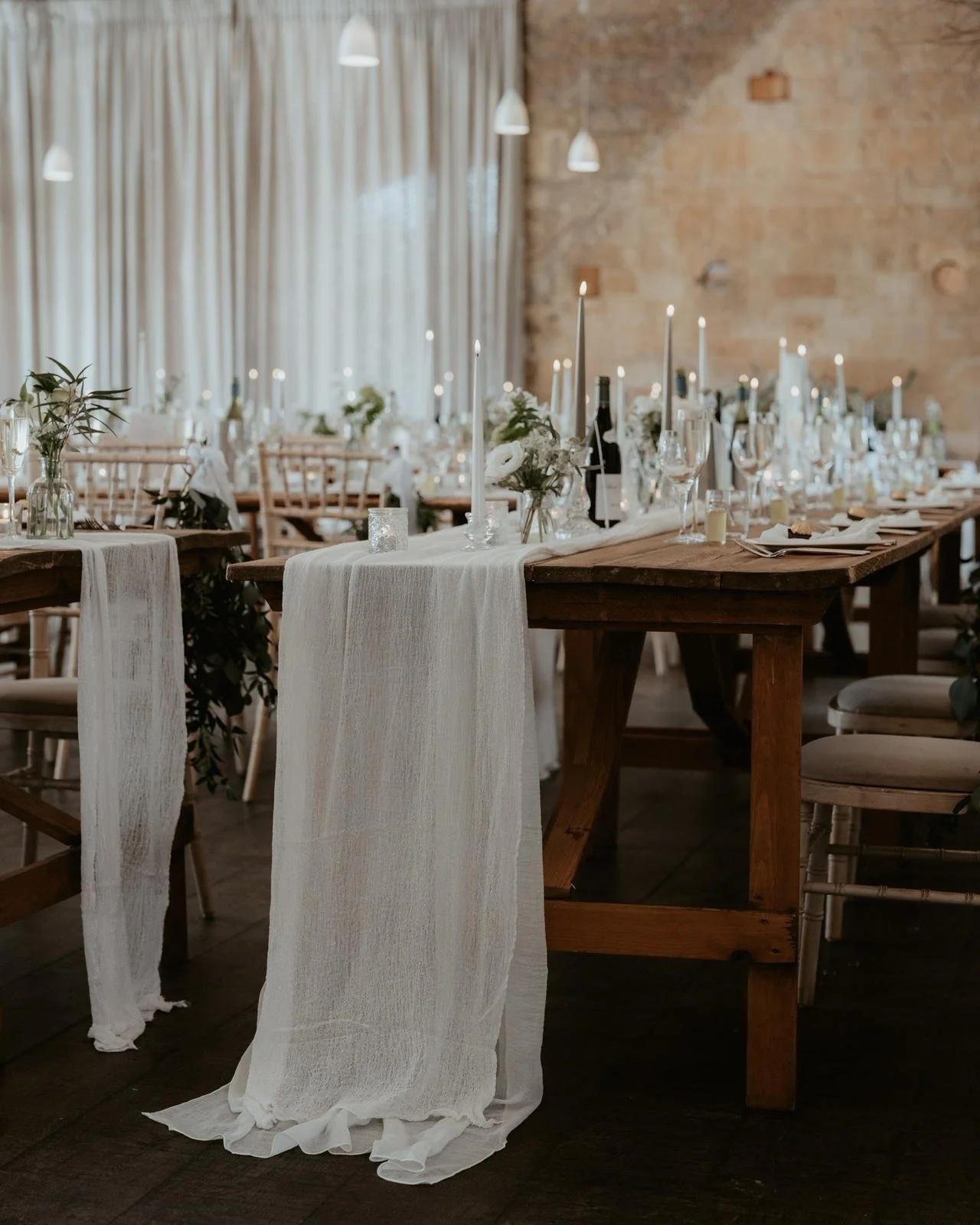 Wedding reception table decorated with white tablecloths, candles, glassware, and floral arrangements in a rustic venue with exposed brick walls.