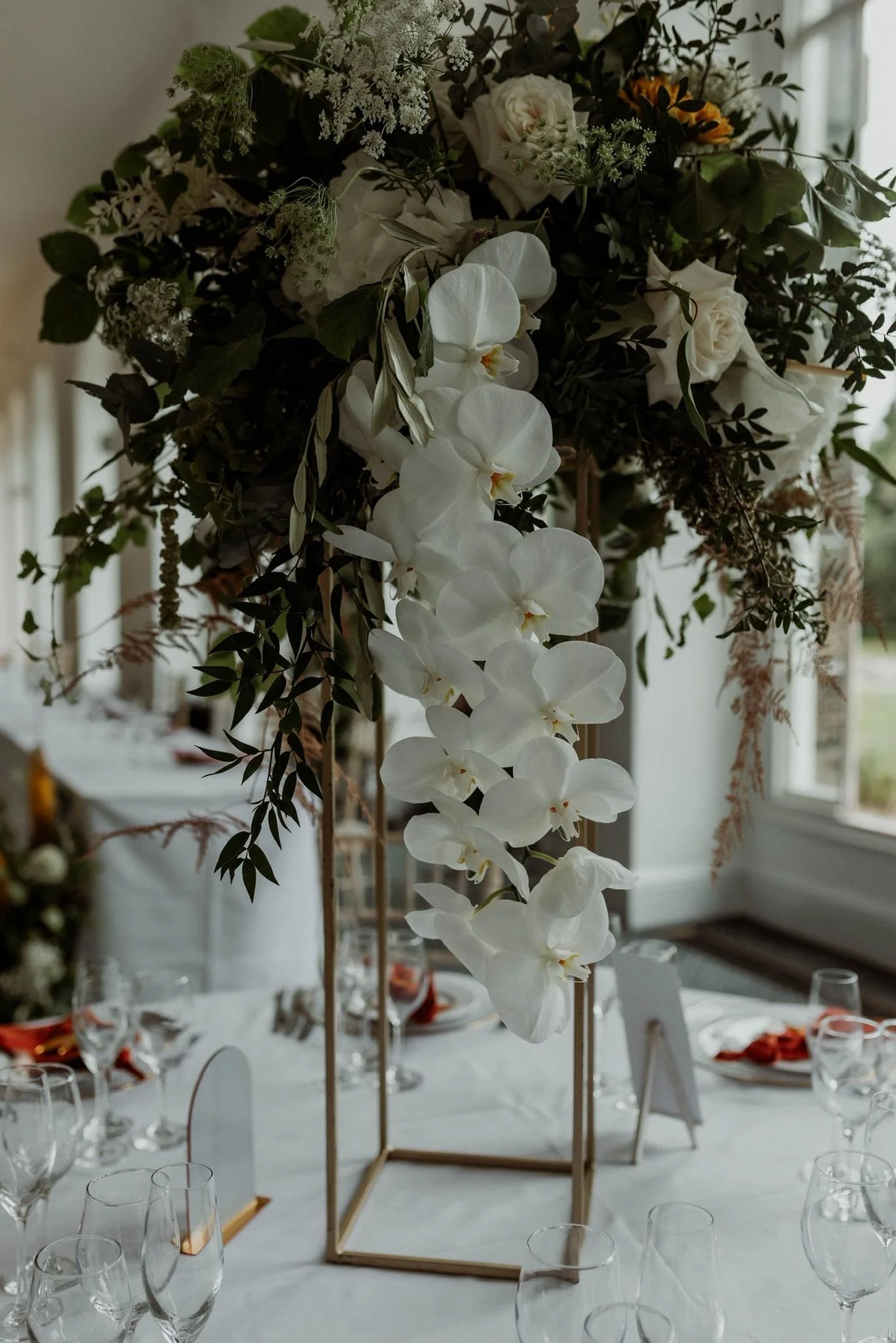 Floral centerpiece with white orchids and roses on a dining table with glasses and table settings in a bright room.