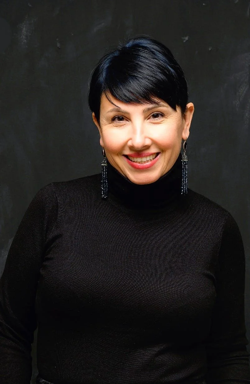 Una donna con capelli corti e neri, indossa un maglione nero a collo alto e orecchini pendenti e sorridente, contro uno sfondo scuro.