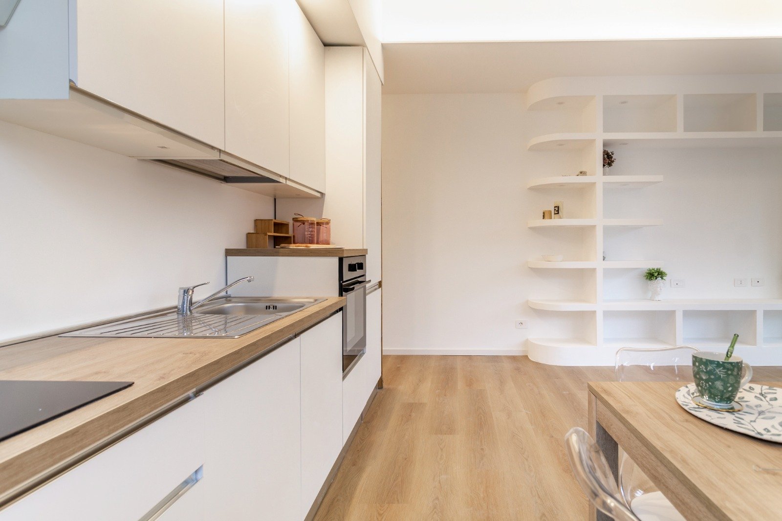 Modern white kitchen with a breakfast bar, four white upholstered chairs, a window seat with colorful cushions, and open shelving with jars and towels, hardwood floors, and recessed lighting.