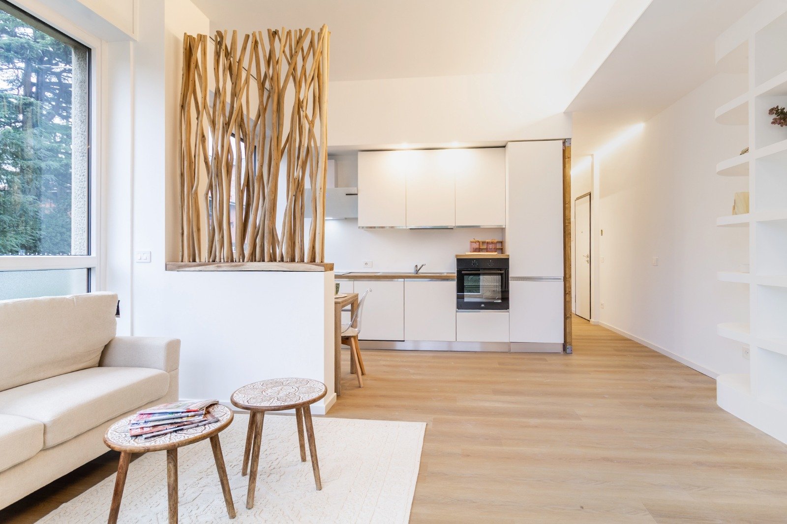 Balcony with yellow cushions, small black table with plants, green artificial grass flooring, white birdcages, overlooking parking lot and green trees under a clear blue sky.