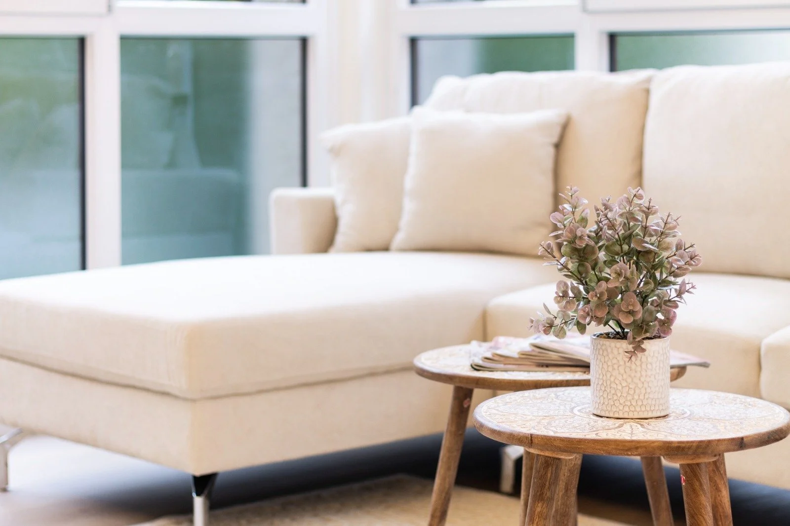 Bright living room with a beige sofa, a wall-mounted bookshelf, a black floor lamp, and large windows with a balcony view of trees.