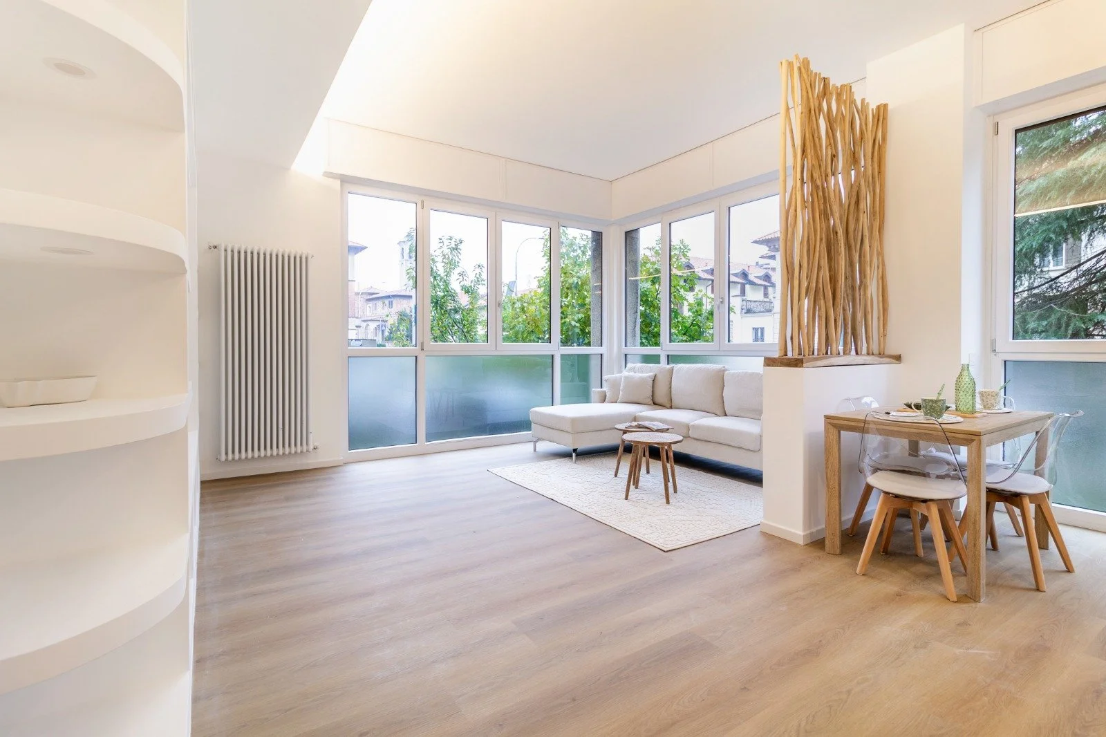 Soggiorno moderno con grandi finestre, divano beige, tappeto chiaro, tavolino e tavolo da pranzo in legno con sedie trasparenti.