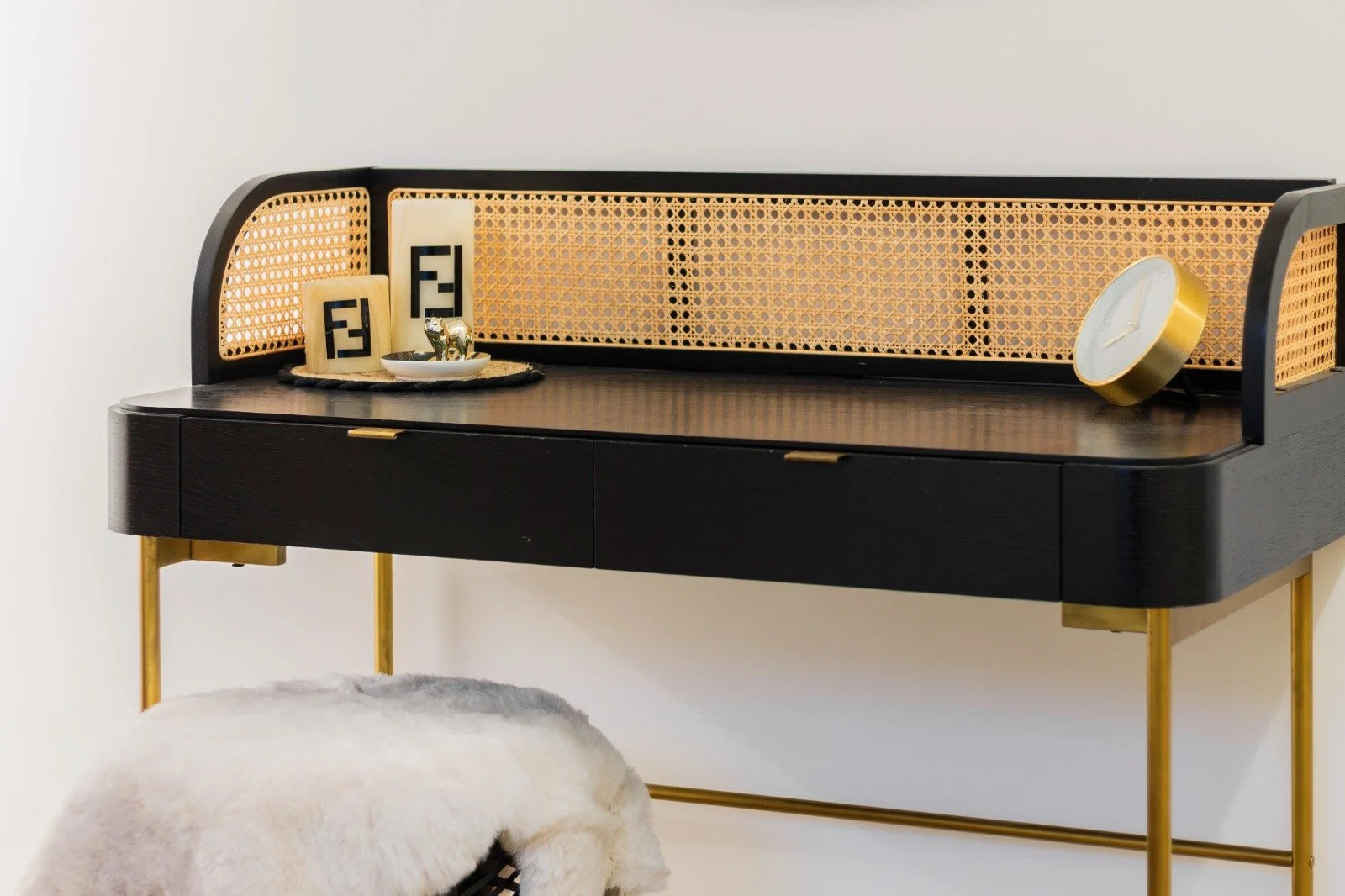 A bedroom with white walls, a single bed with white bedding and floral pillow, pink blanket, wooden floor, and a fluffy gray rug. There is a white shelf with a photo frame and a small stuffed animal. An open door shows a living area with a black floor lamp and glass doors leading outside.