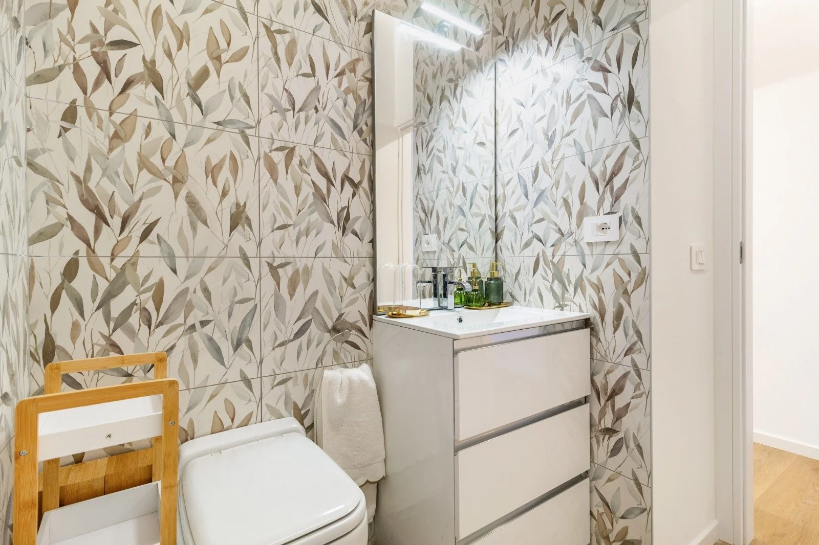 A table with a decorative face-shaped white vase holding a green plant, a small green pot with a succulent, and two colorful pillows with animal patterns by a window, with a yellow patterned lamp nearby.