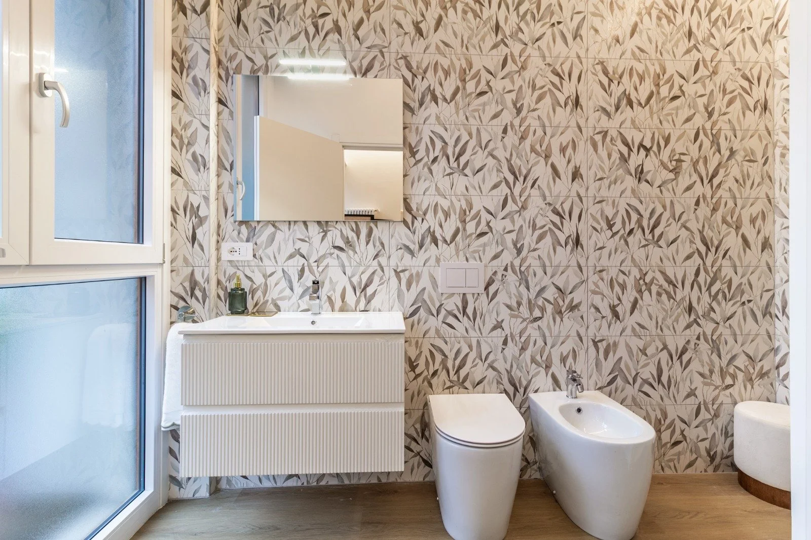 A modern bathroom with a terrazzo shower, a white bidet and toilet, a green vanity with a white sink, a large mirror, and a window with a wooden sill.