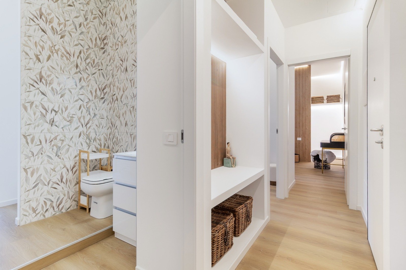 Corner of a room with a wooden windowsill, a lamp with a patterned lampshade, a window showing green trees outside, and two decorative pillows with orange, white, and brown patterns.