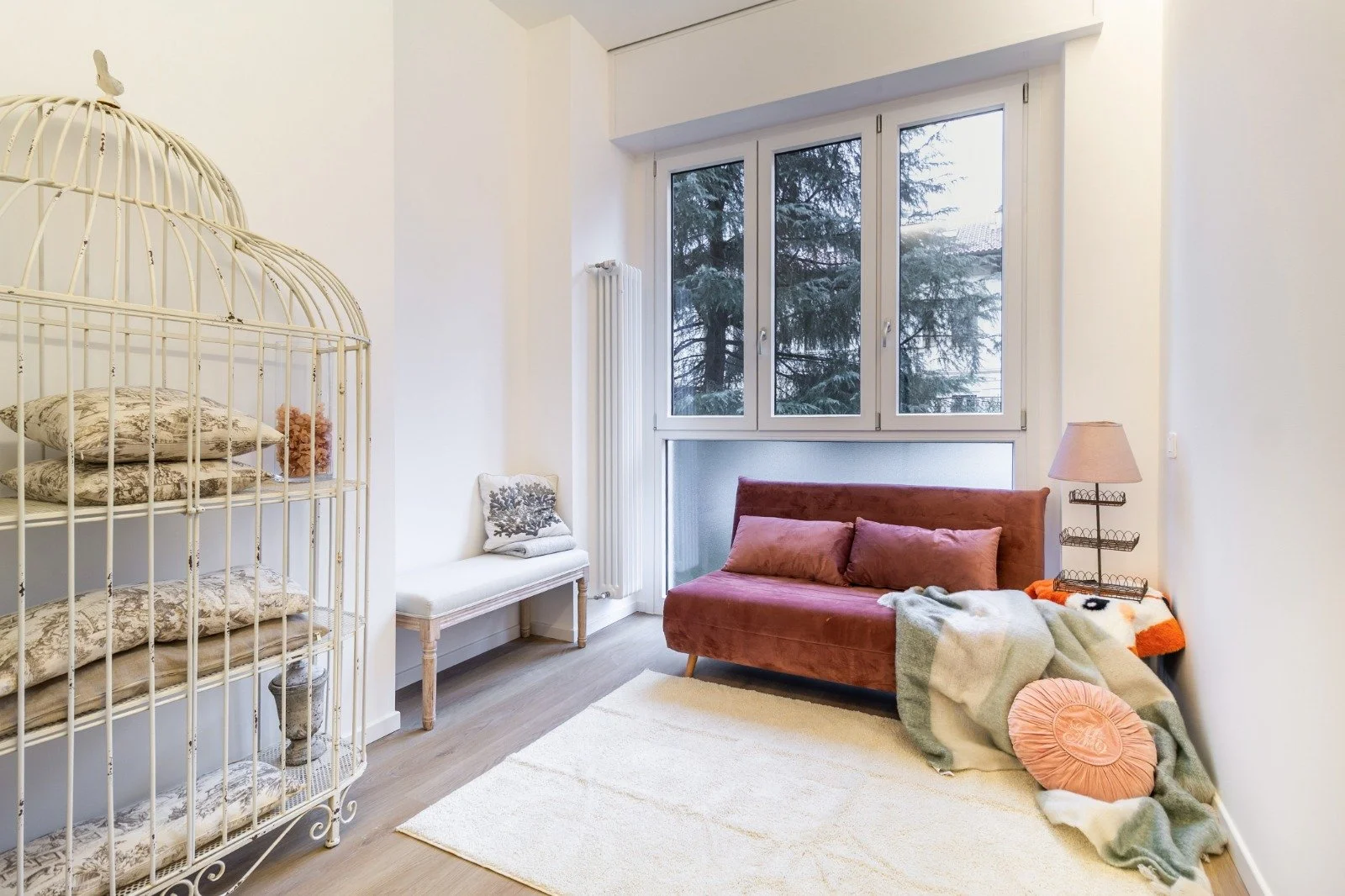 Modern dining room with large window, white walls, and light wood floor. Features a round table with three beige chairs, a pink rug, and minimalist white shelves with decorative items.