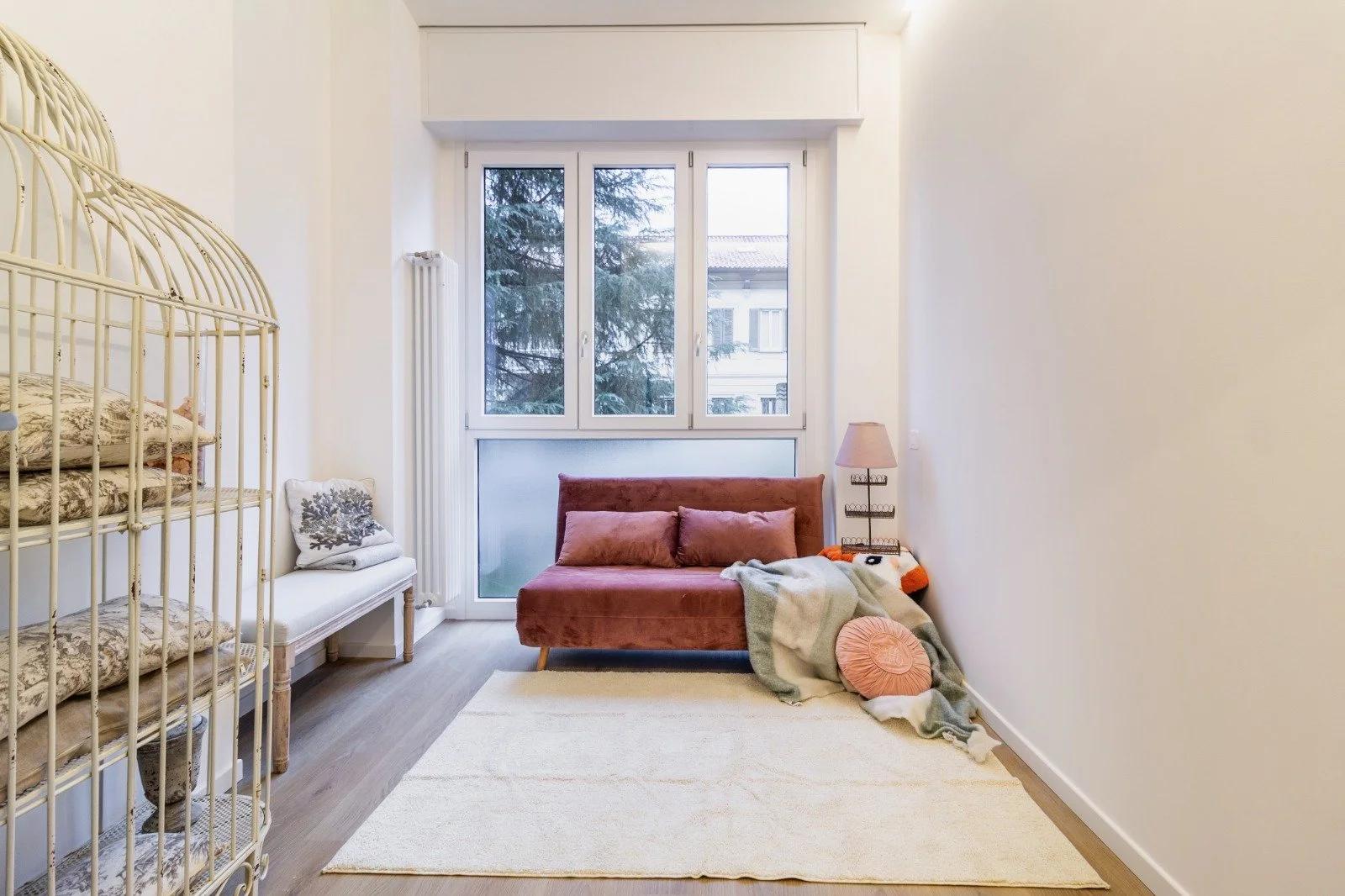 Modern living room with light-colored walls, wooden flooring, a beige sofa with a throw blanket, a tall black floor lamp, a shelf with decor items, and a large window with a balcony view.