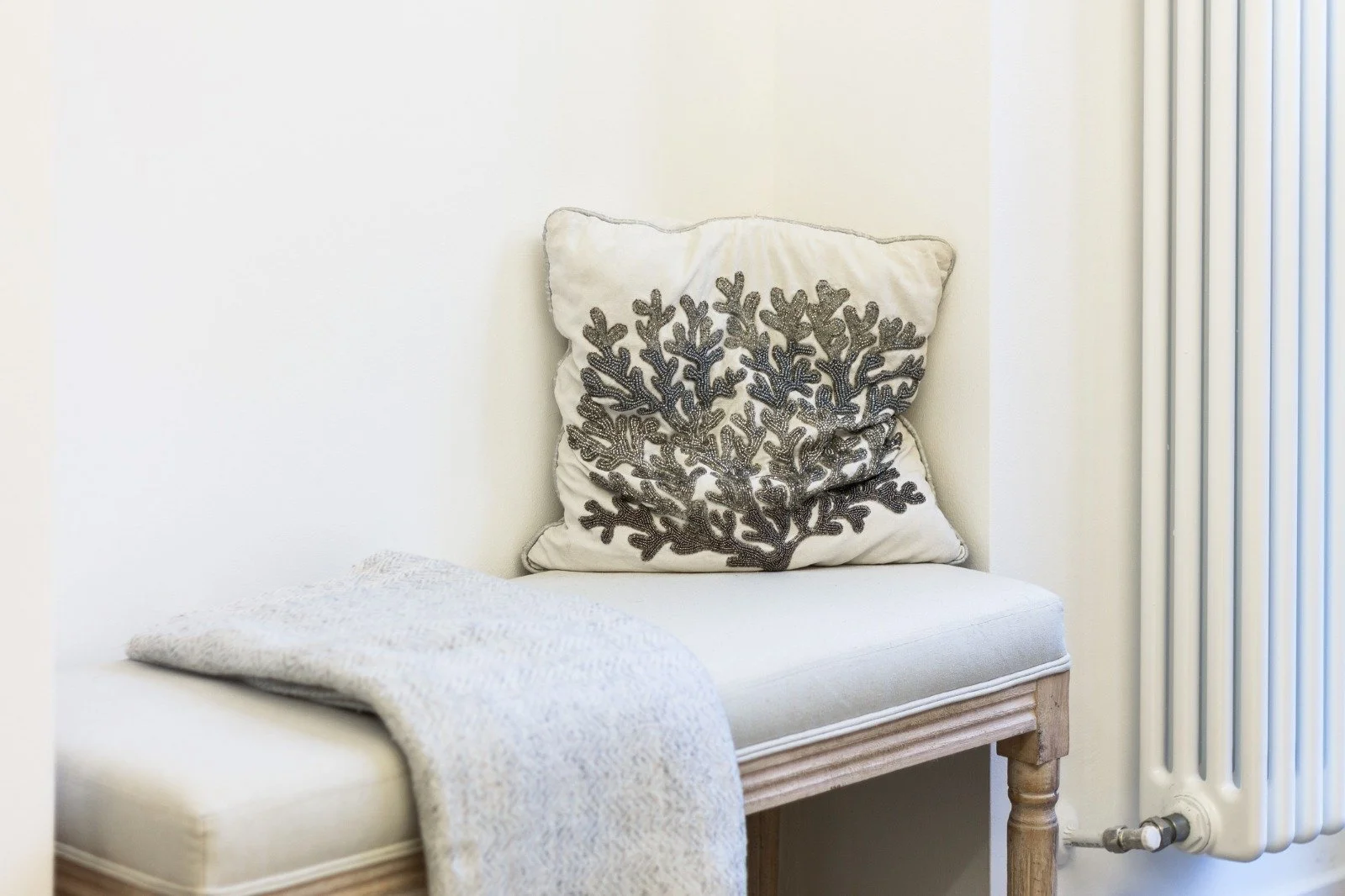 A modern dining room with a white round table, a beige upholstered chair, and a gold base. On the table, there is a round gold tray with two decorative candles featuring black geometric symbols. A window with greenery outside is visible in the background.