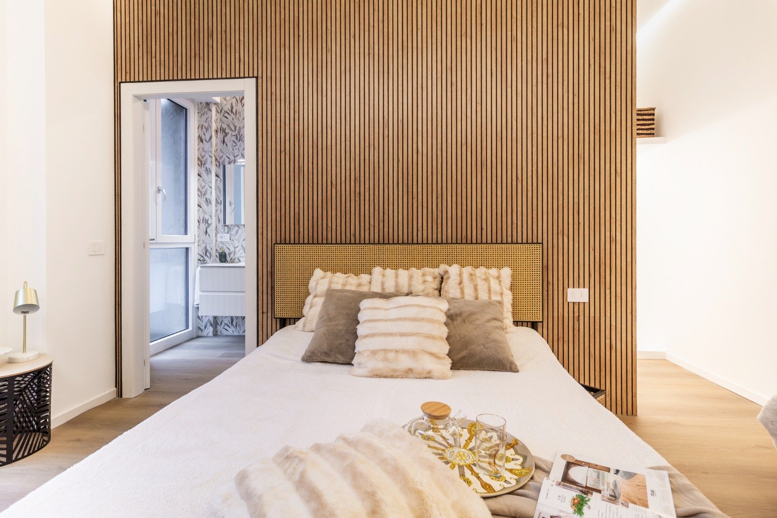 Bright bedroom with two beds, one with floral pillows and pink blanket, the other with beige pillows and pink blanket, a balcony door showing trees outside, a white rug, a small white ottoman, and minimal decor.