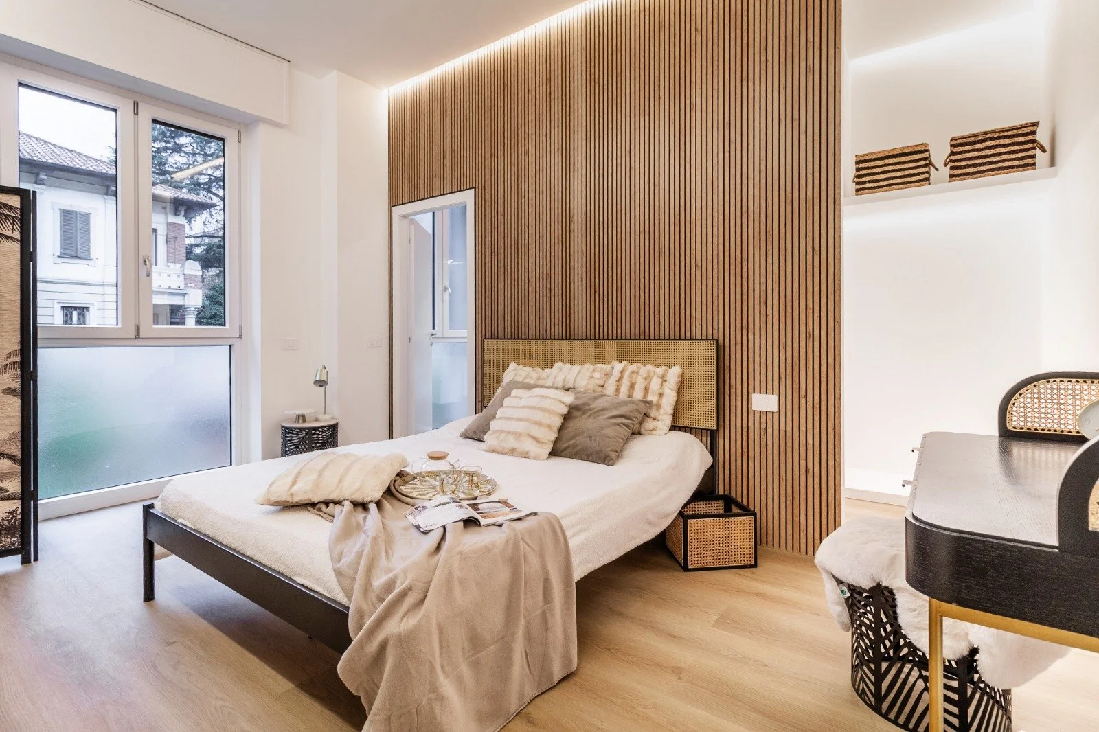 Modern living room with white walls, a round marble table, two white candles with black logo, a beige lounge chair, and multiple white and gold shelves decorated with books and plants.