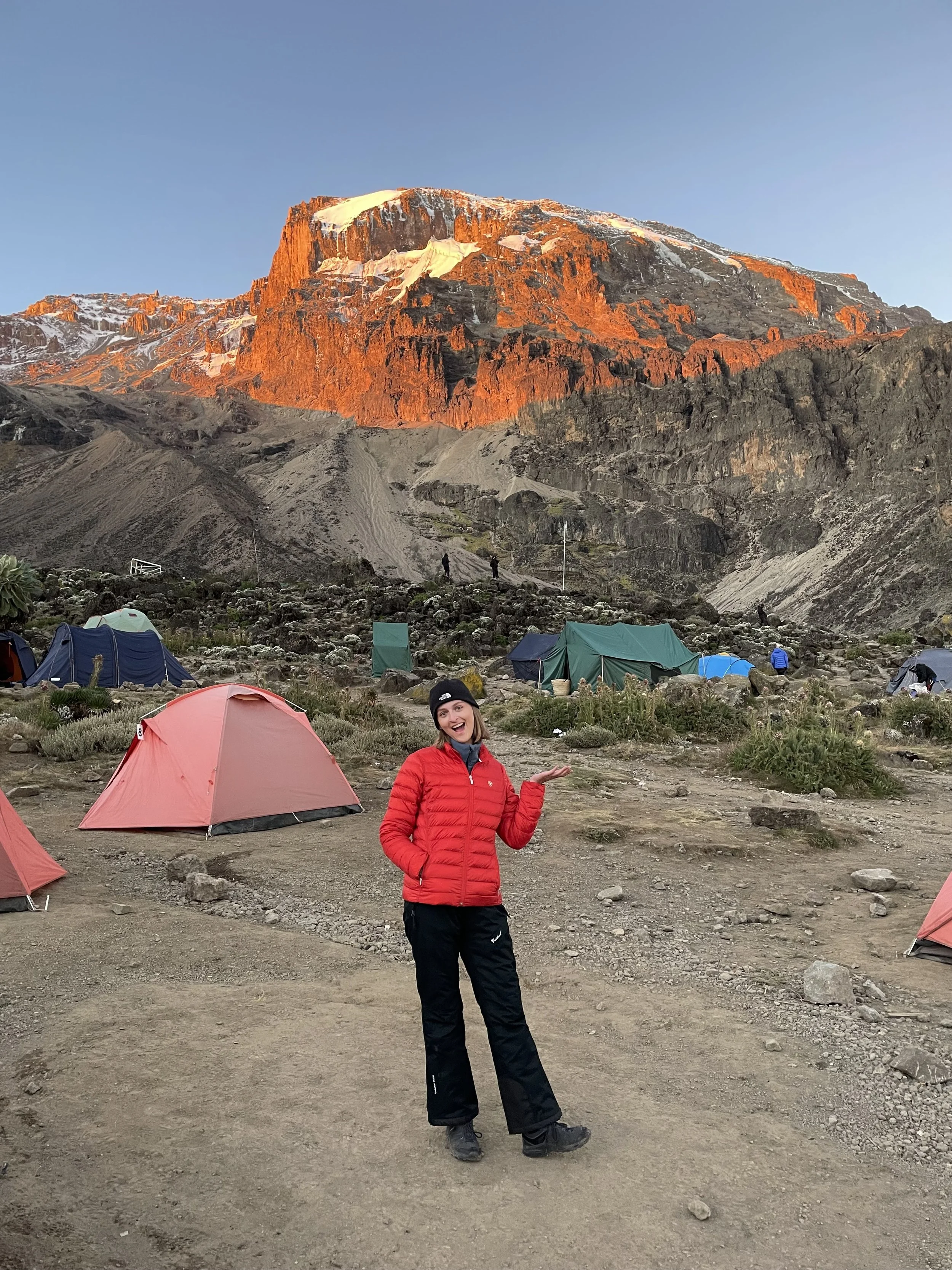 A woman smiling and pointing to the right, standing in front of a campsite with tents, with a mountain illuminated by the setting or rising sun in the background.