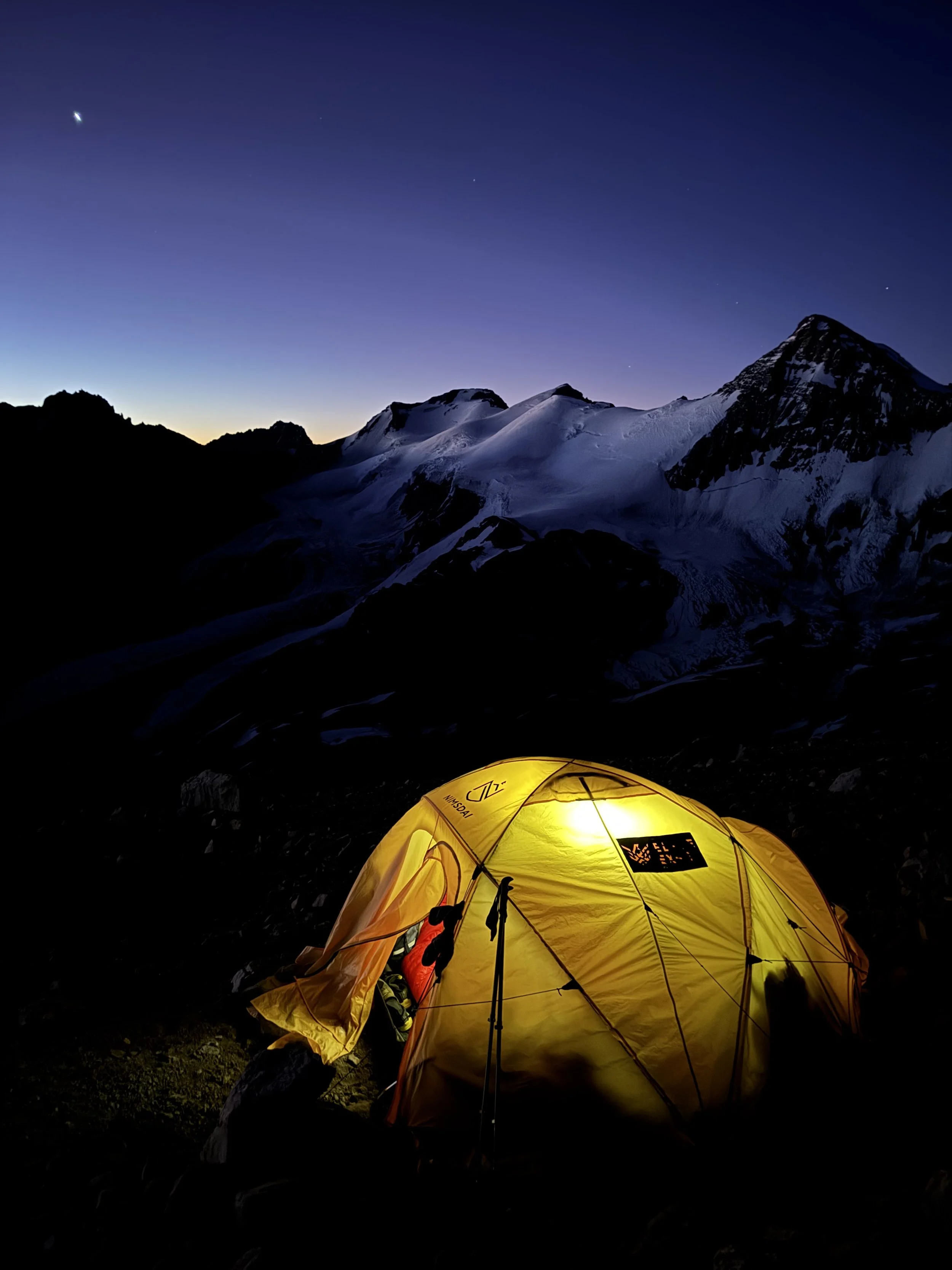 A yellow tent lit from within sits on rocky terrain at night with snow-covered mountains in the background and a dark sky with stars above.