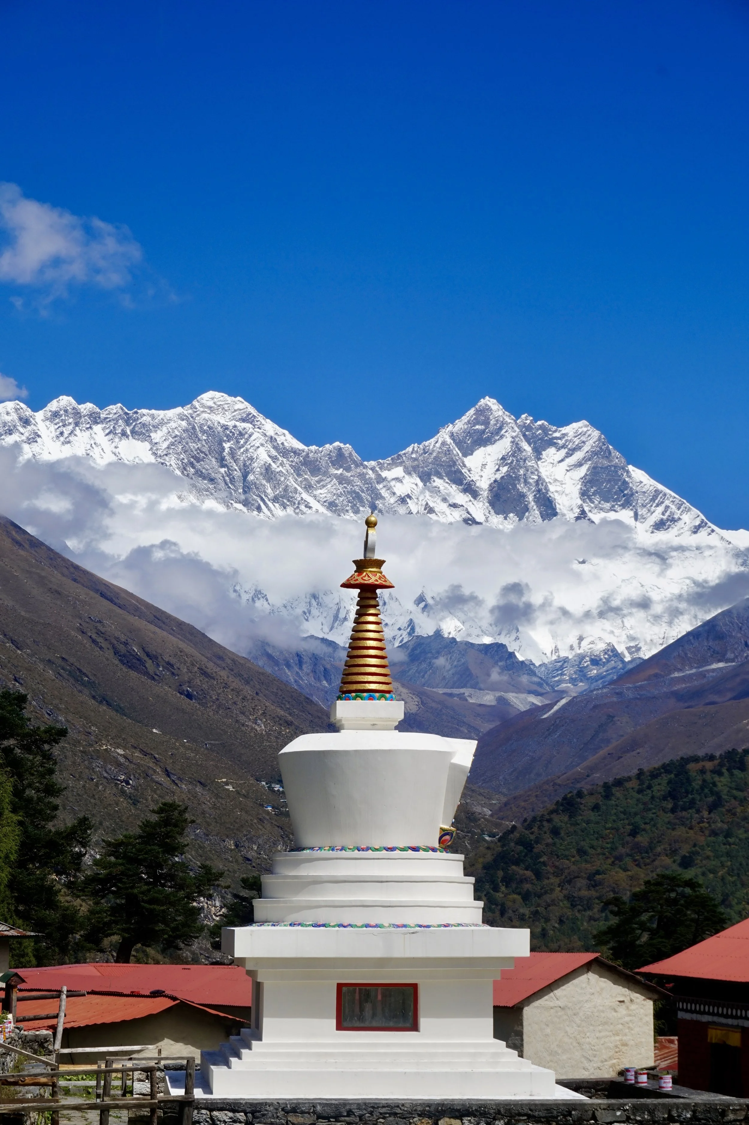 A white Buddhist stupa with a decorative top, set against a mountainous landscape with snow-capped peaks and a clear blue sky.