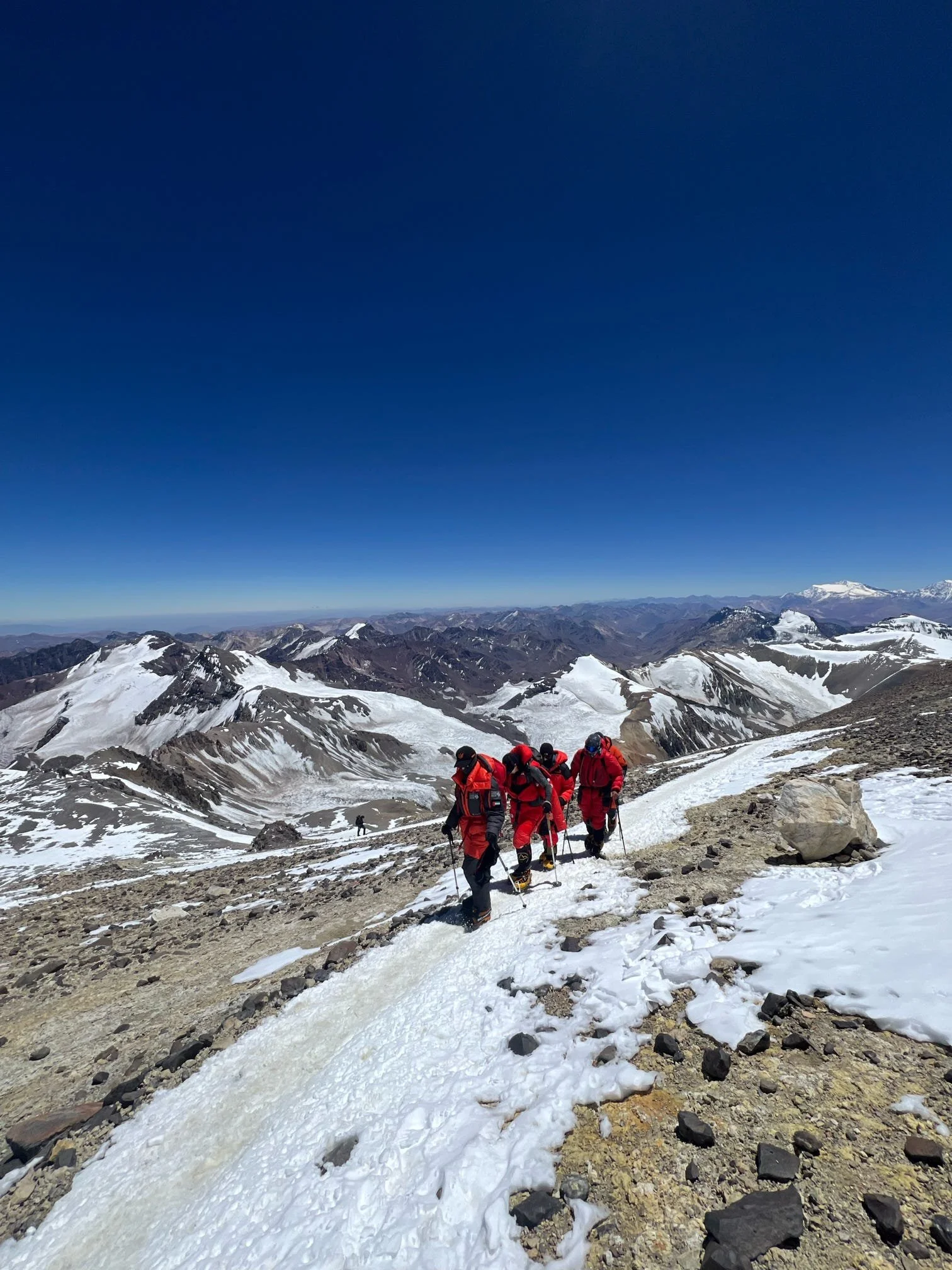A group of hikers dressed in red jackets and black gear climbing snowy mountain terrain under a clear blue sky.