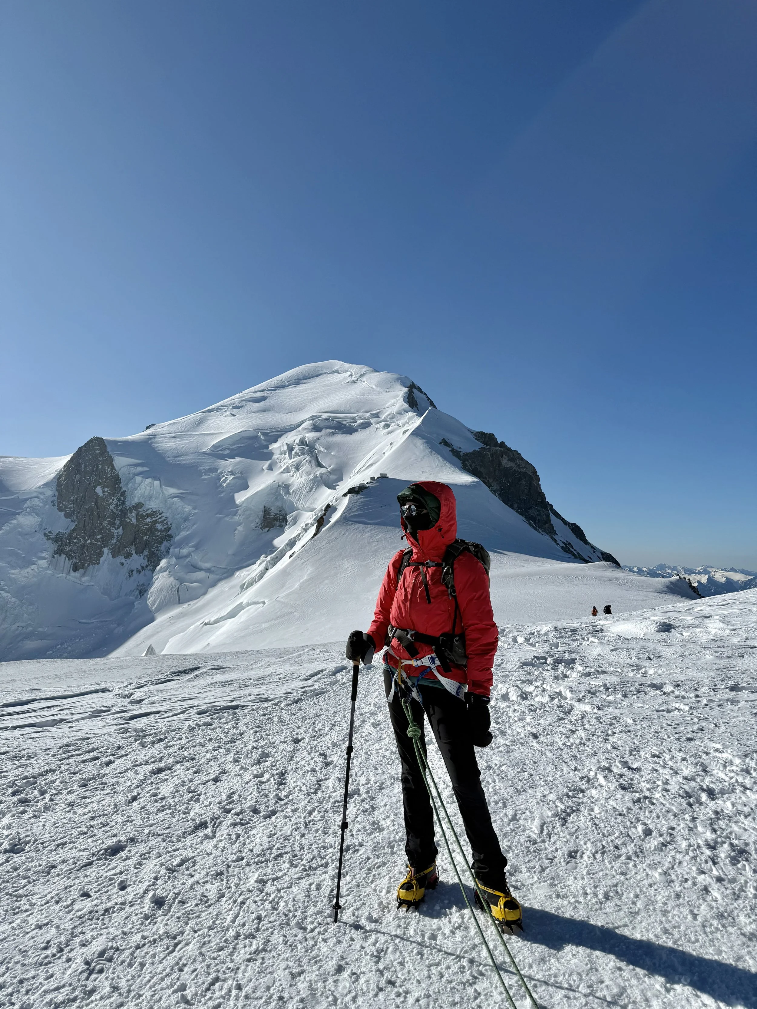 A person in red winter clothing with ski gear standing on snow-covered mountain, with a snow-capped mountain peak in the background under a clear blue sky.