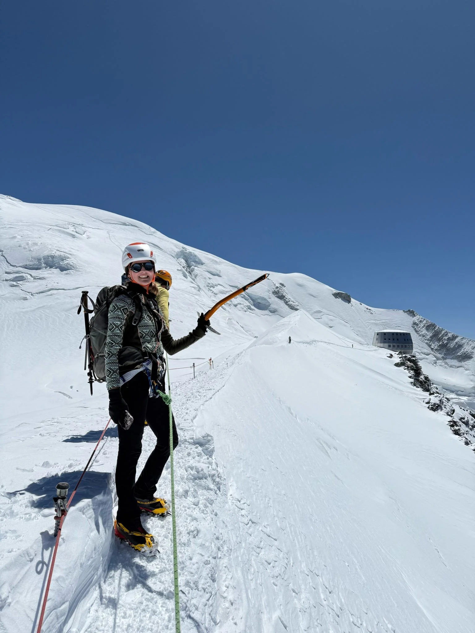 A woman in mountaineering gear and yellow snowshoes standing on snow, holding an ice axe, with a snowy mountain and clear blue sky in the background.