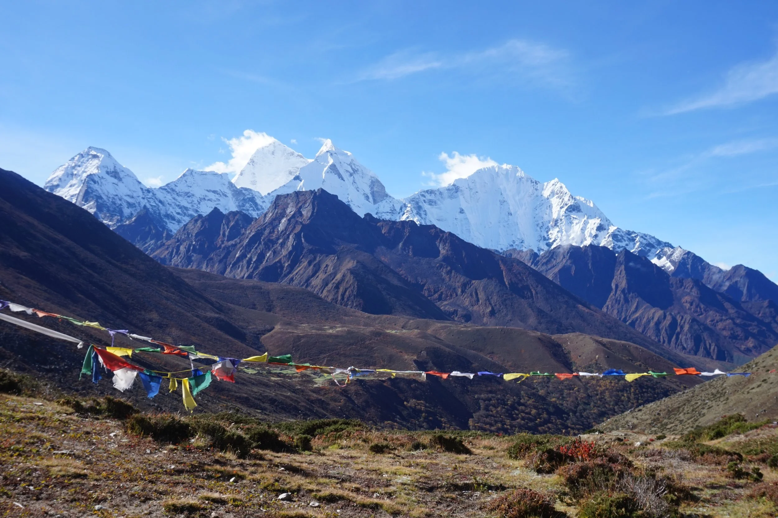 Snow-capped mountains in the background with colorful prayer flags in the foreground, blue sky with some clouds.