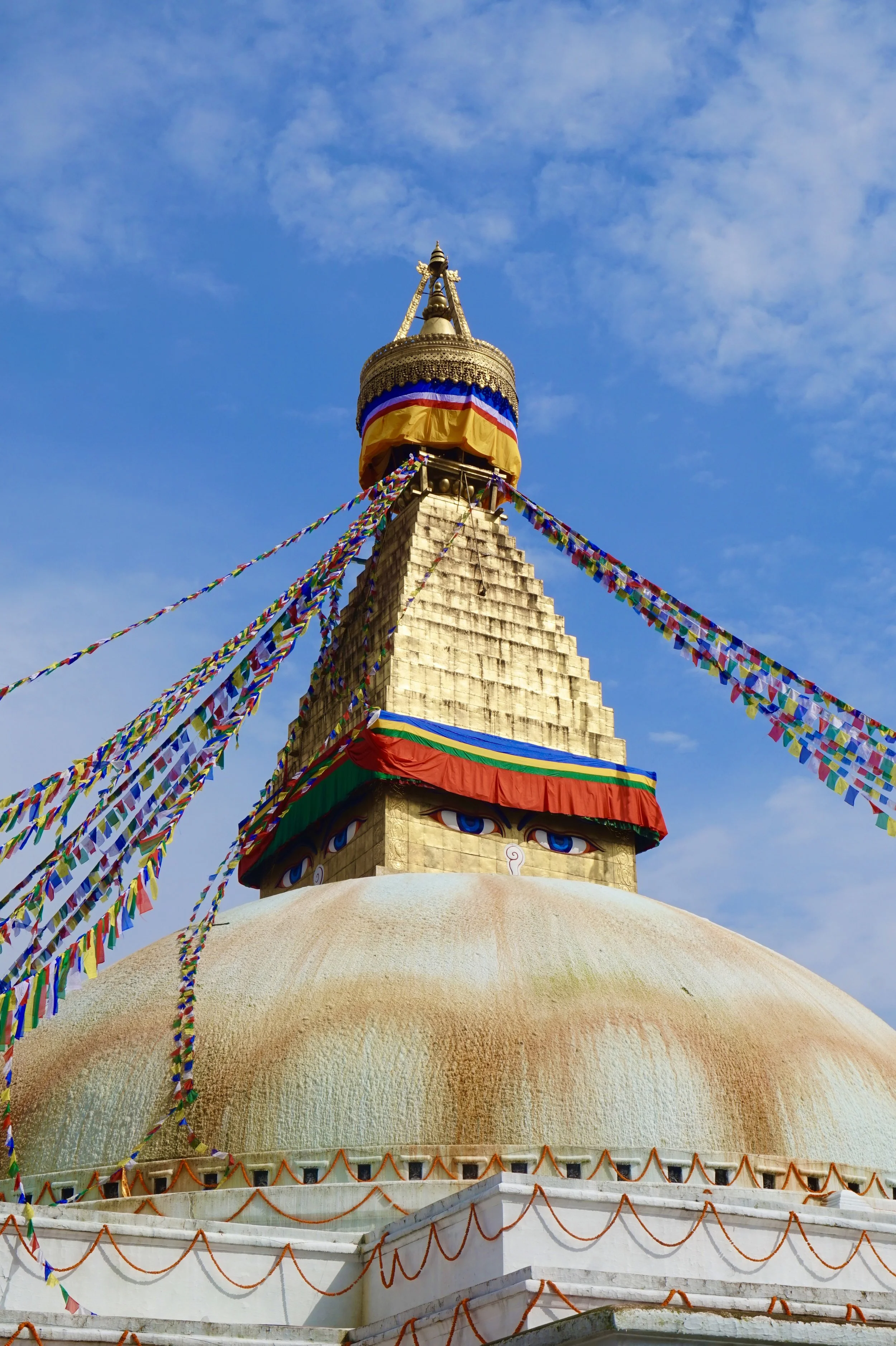 A large white Buddhist stupa with a golden spire, decorated with colorful prayer flags, in front of a blue sky with some clouds.