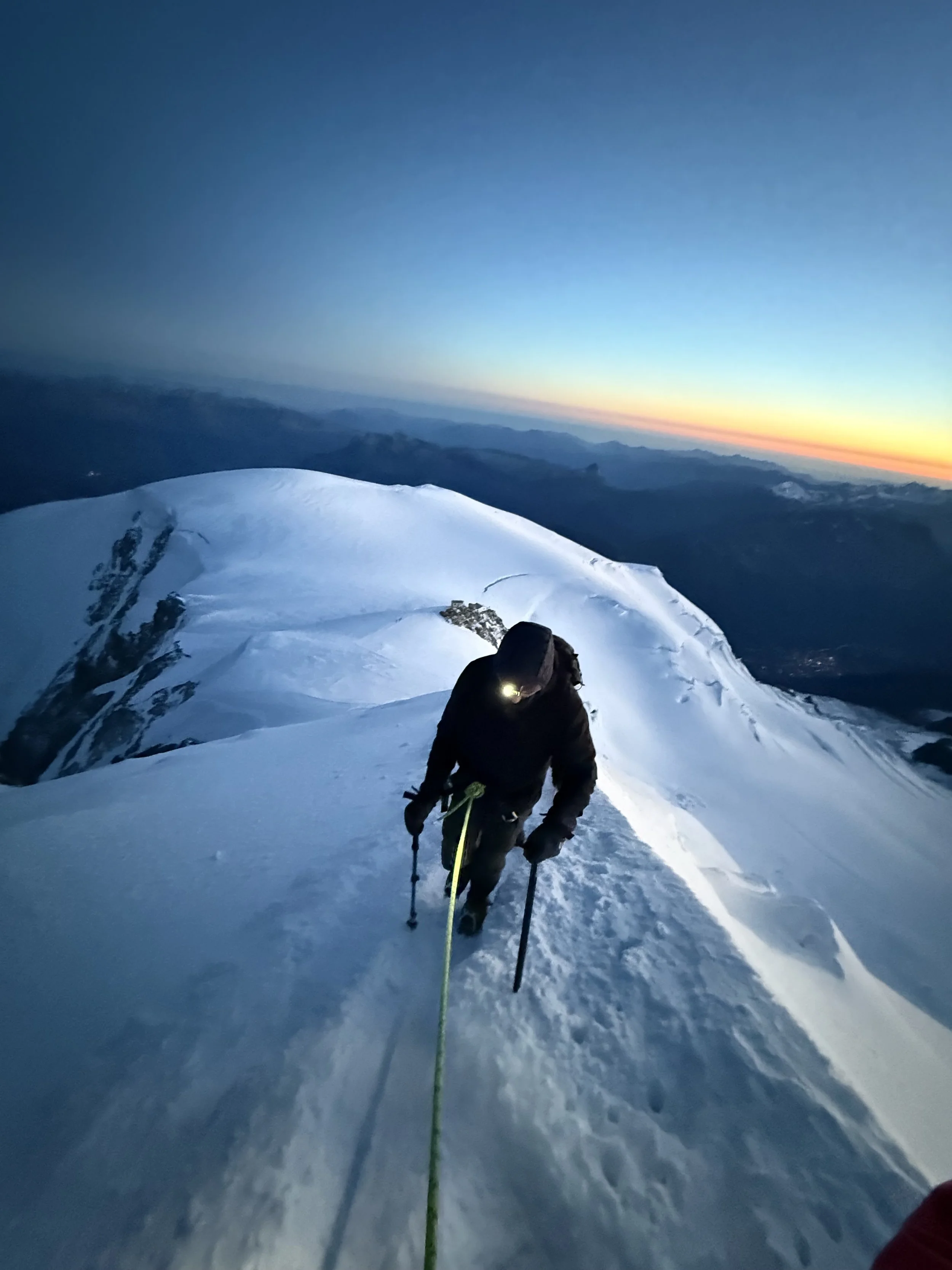 A mountain climber ascending a snow-covered mountain ridge at dawn or dusk, with a distant horizon and a colorful sky