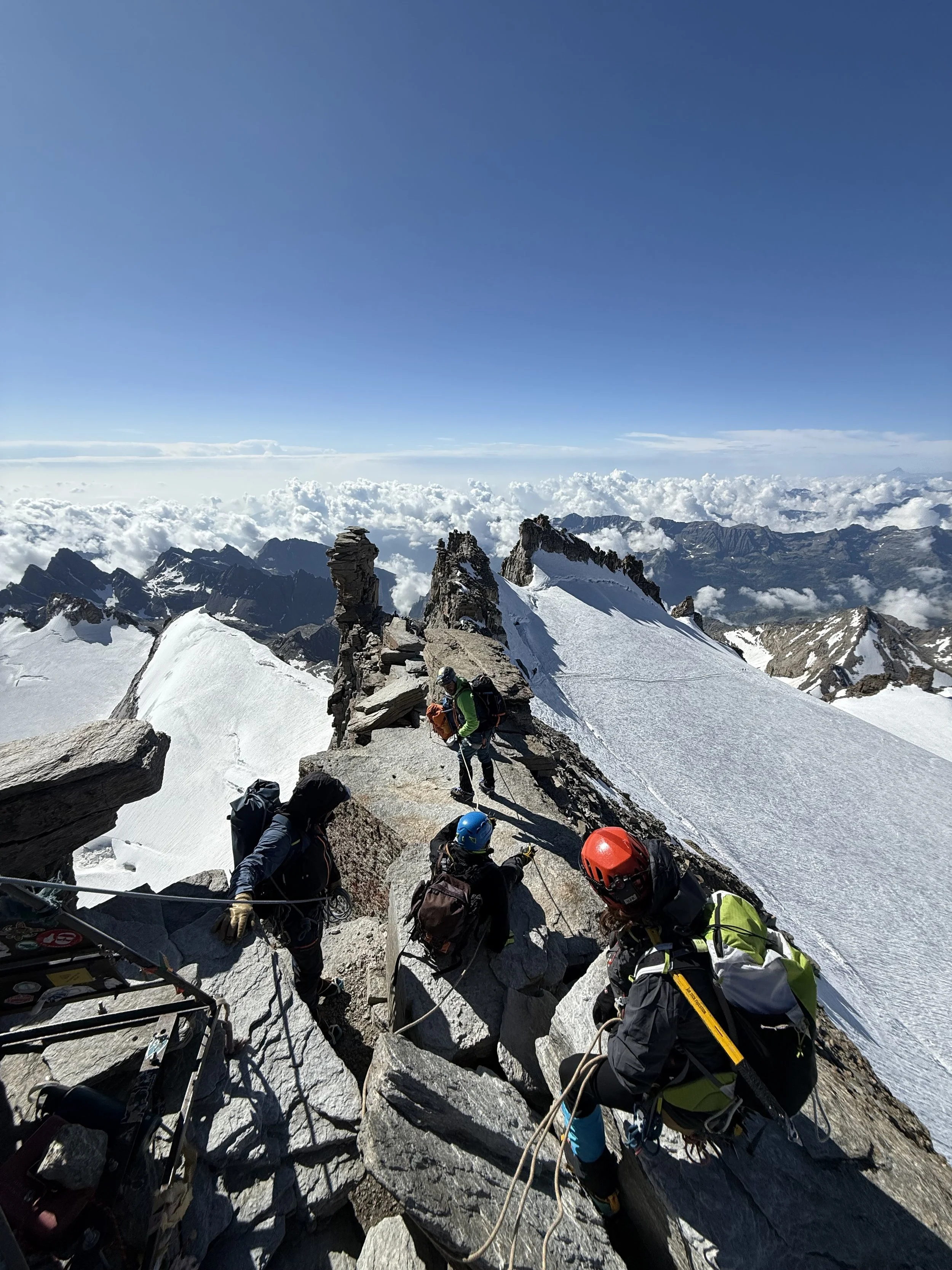 Mountaineers climbing a rocky, snow-covered mountain ridge at high altitude with a background of clouds and mountain peaks under a clear blue sky.