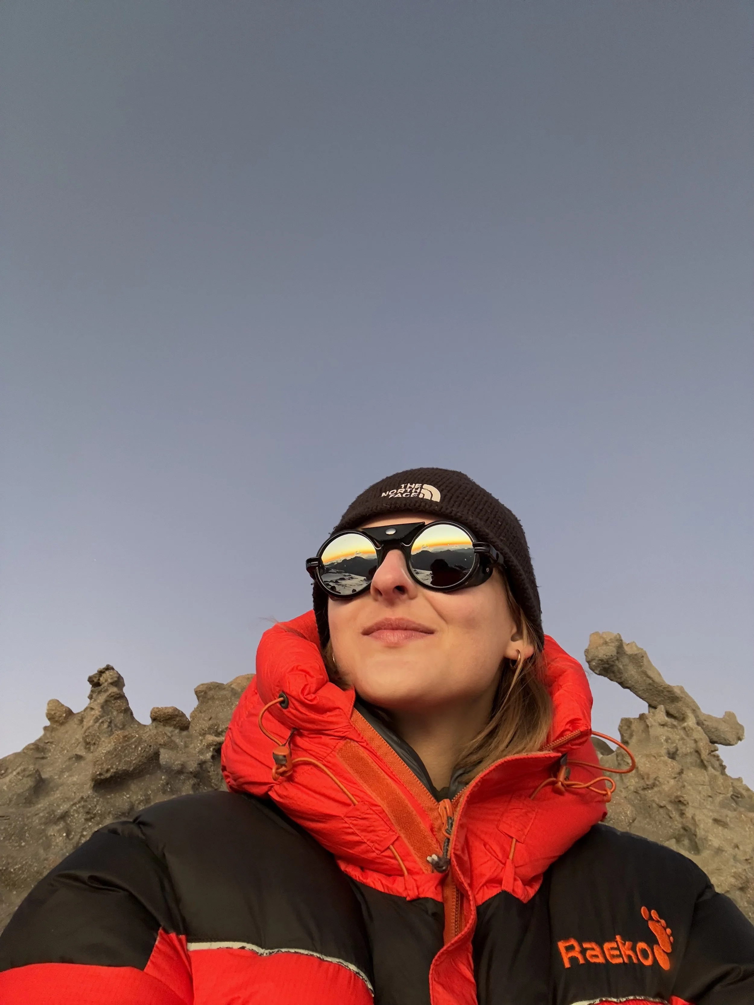 A woman in outdoor gear, wearing sunglasses, a black beanie, and a red and black jacket, standing in front of rocky terrain with a clear sky behind her.