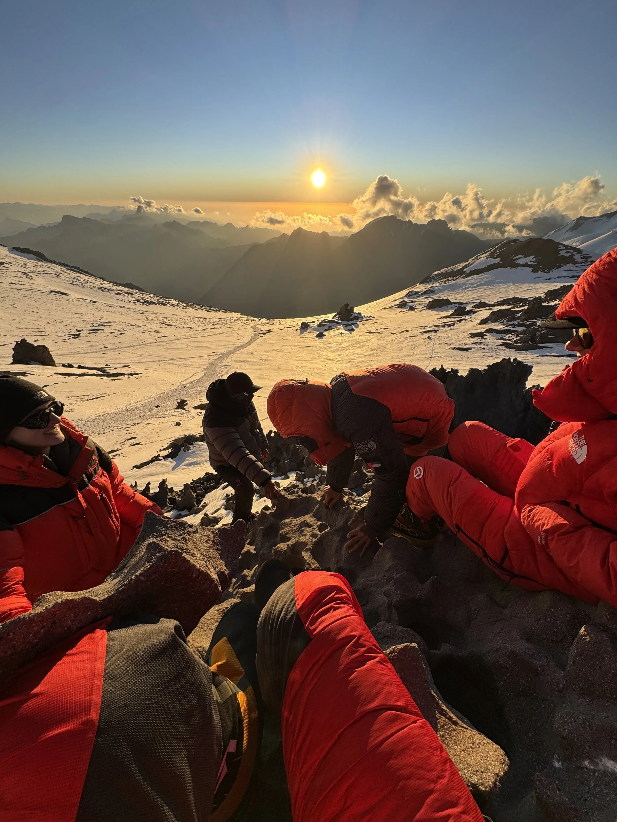 Group of climbers in red jackets resting on snowy mountain ridge during sunset.