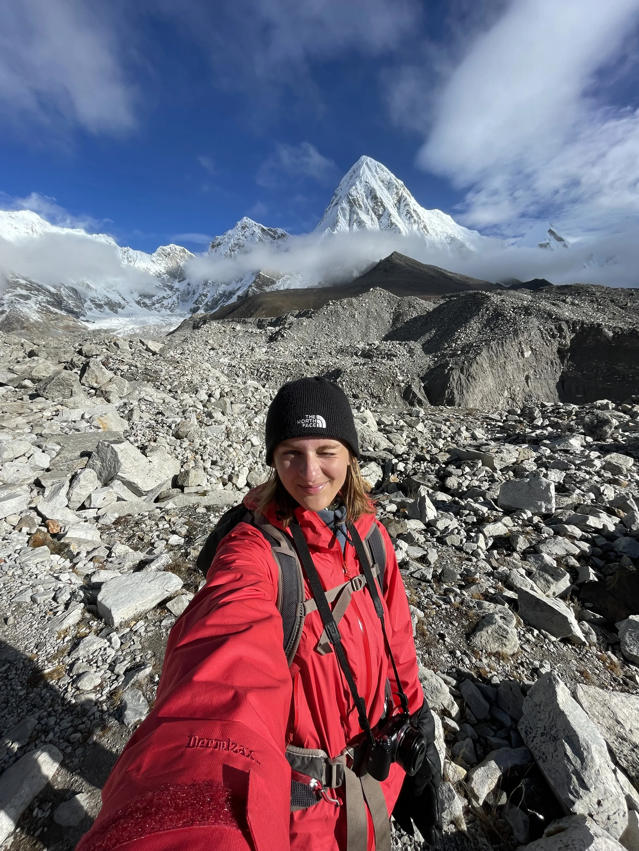 A woman in a red jacket and black beanie taking a selfie on rocky terrain with snow-capped mountains in the background under a blue sky.