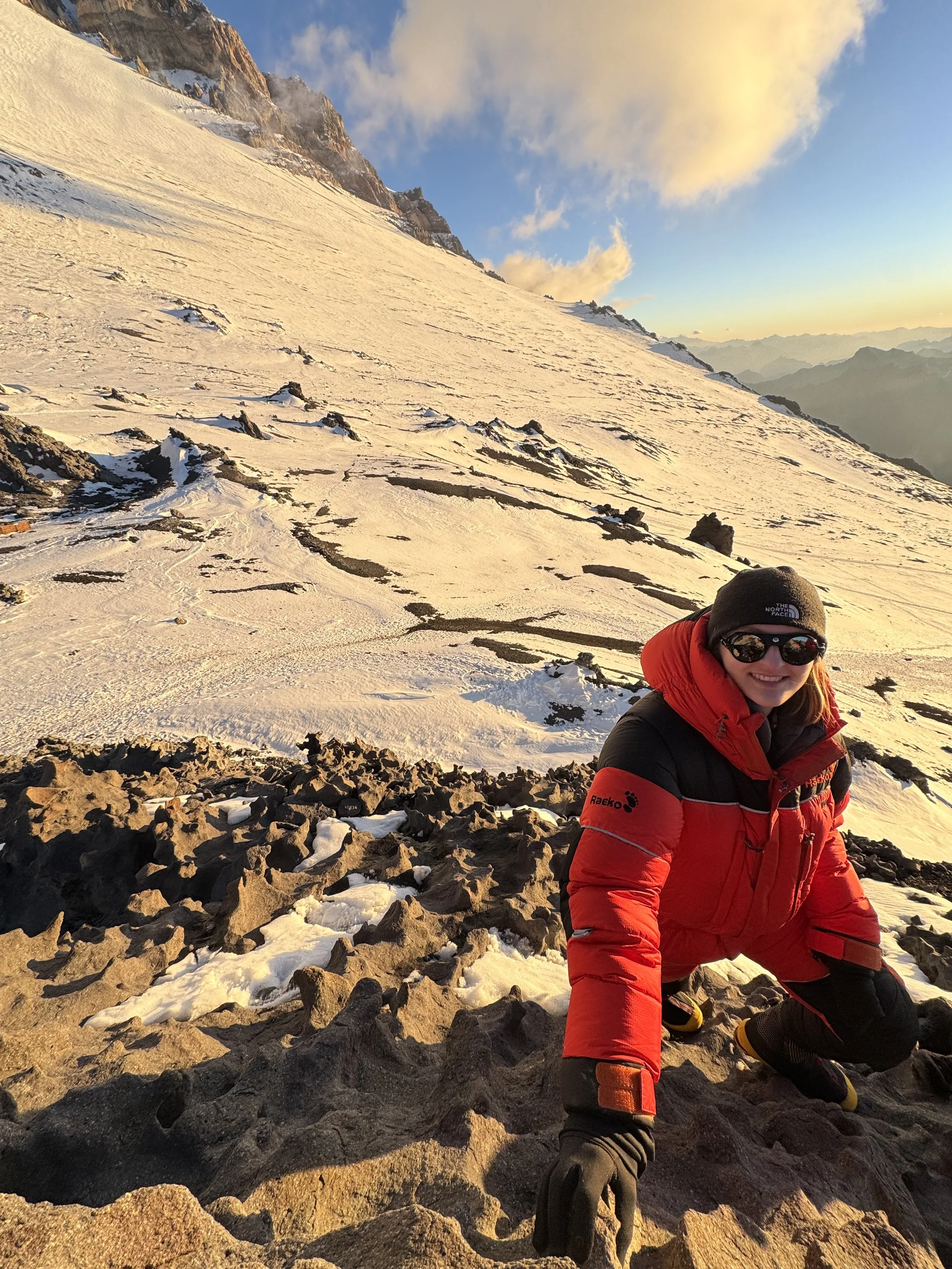 A person in red outdoor gear smiling while kneeling on a rocky, snow-covered mountain slope during sunset with snow-capped peaks in the background.