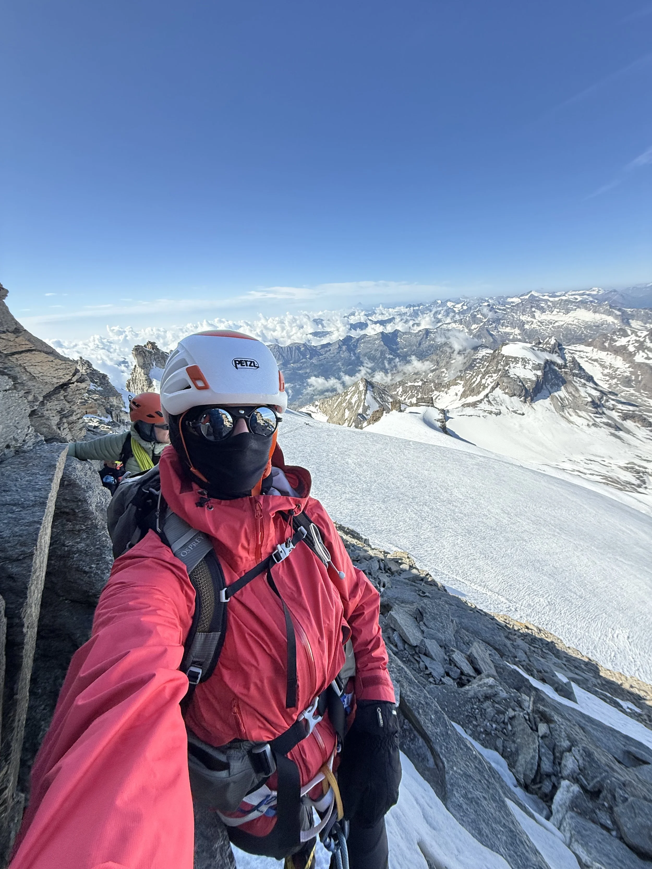 A climber in red gear and a white helmet taking a selfie on a snowy mountain ridge with a mountain range in the background.