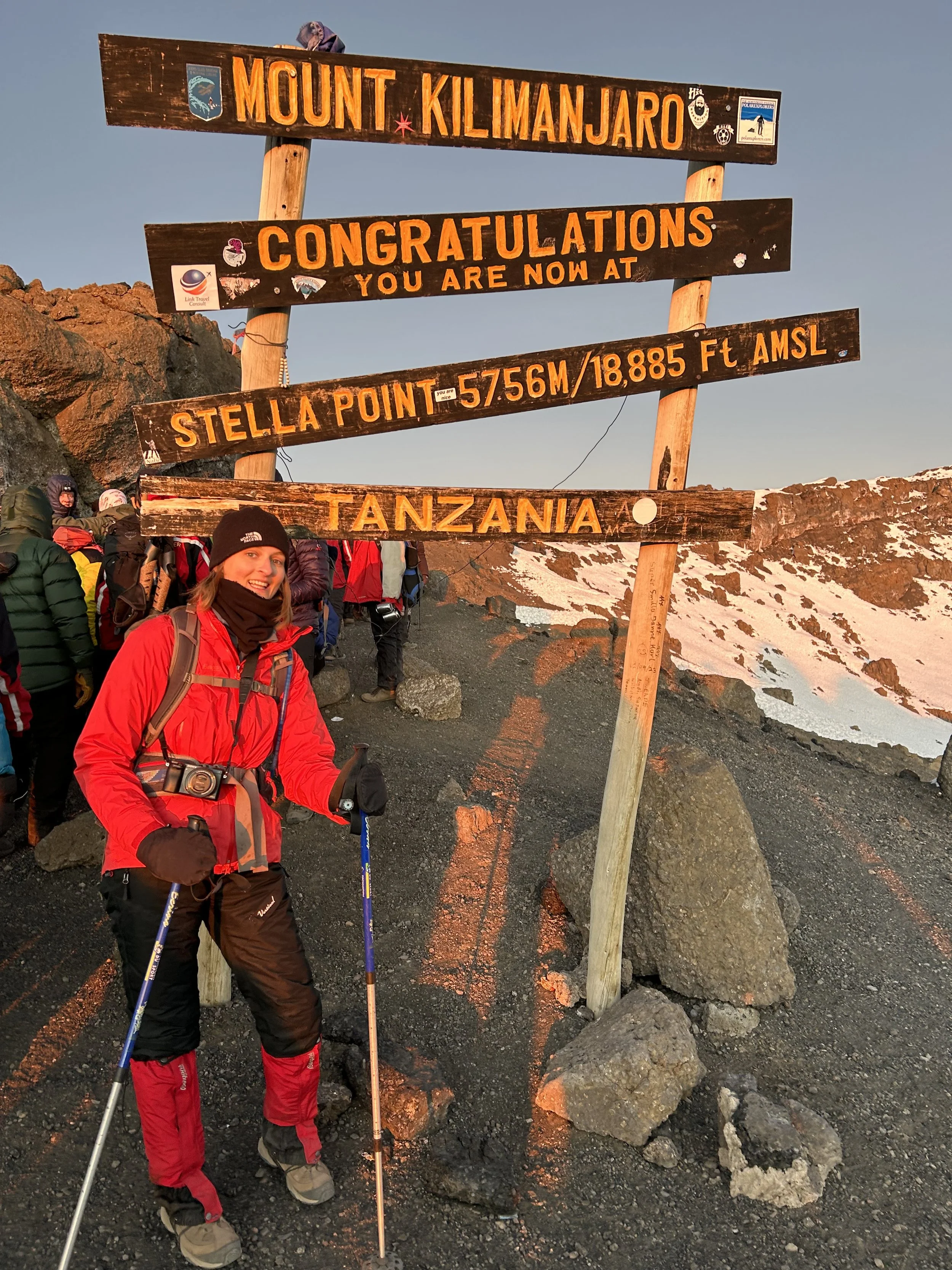 A woman dressed in red and black outdoor gear, holding trekking poles, standing next to a sign at Mount Kilimanjaro indicating the summit at 5,756 meters or 18,885 feet above sea level in Tanzania during sunset.