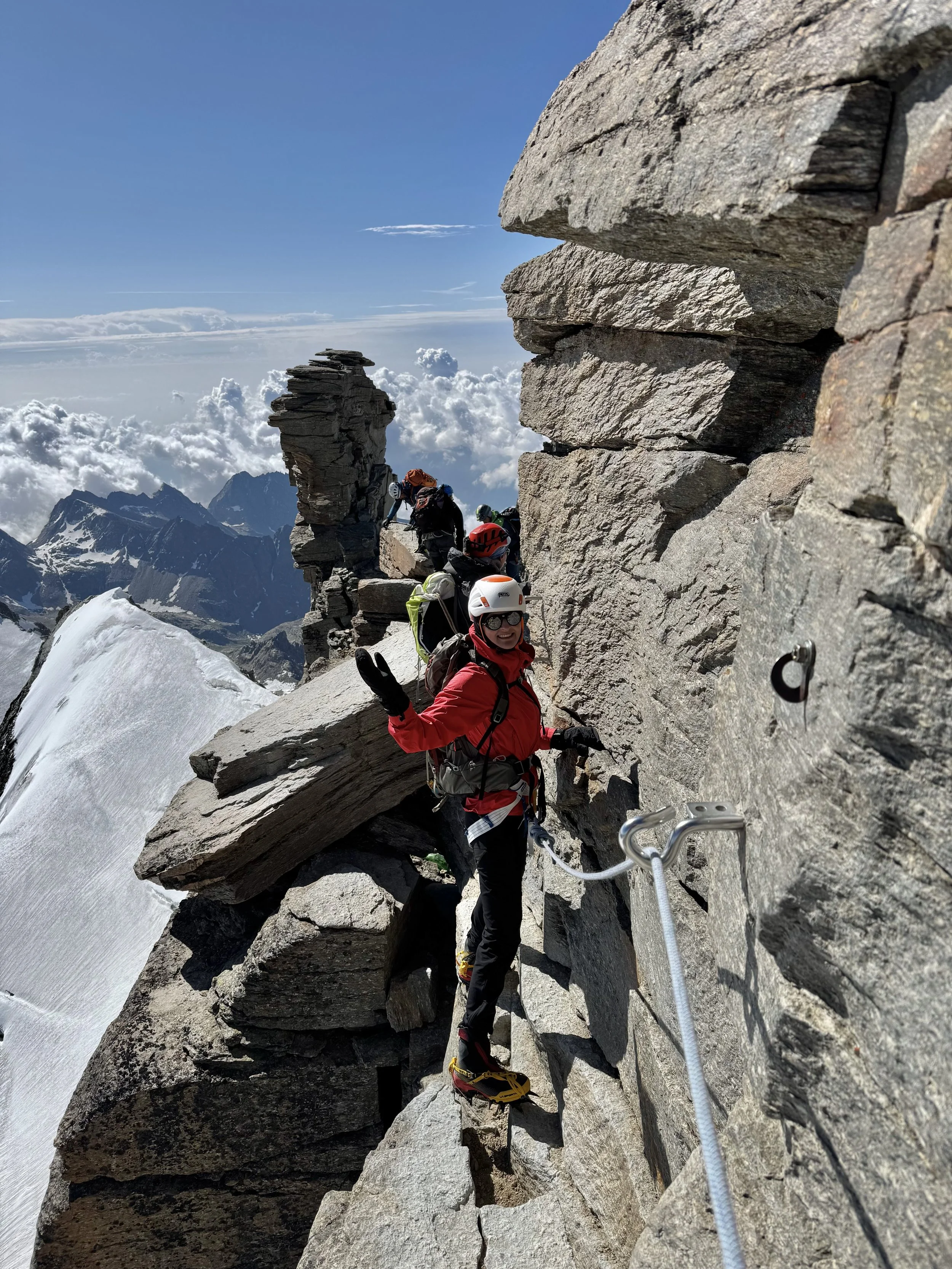 Group of climbers ascending a mountain ridge with metal safety cables, surrounded by rocky cliffs and snow, with a backdrop of mountain peaks and partly cloudy sky.