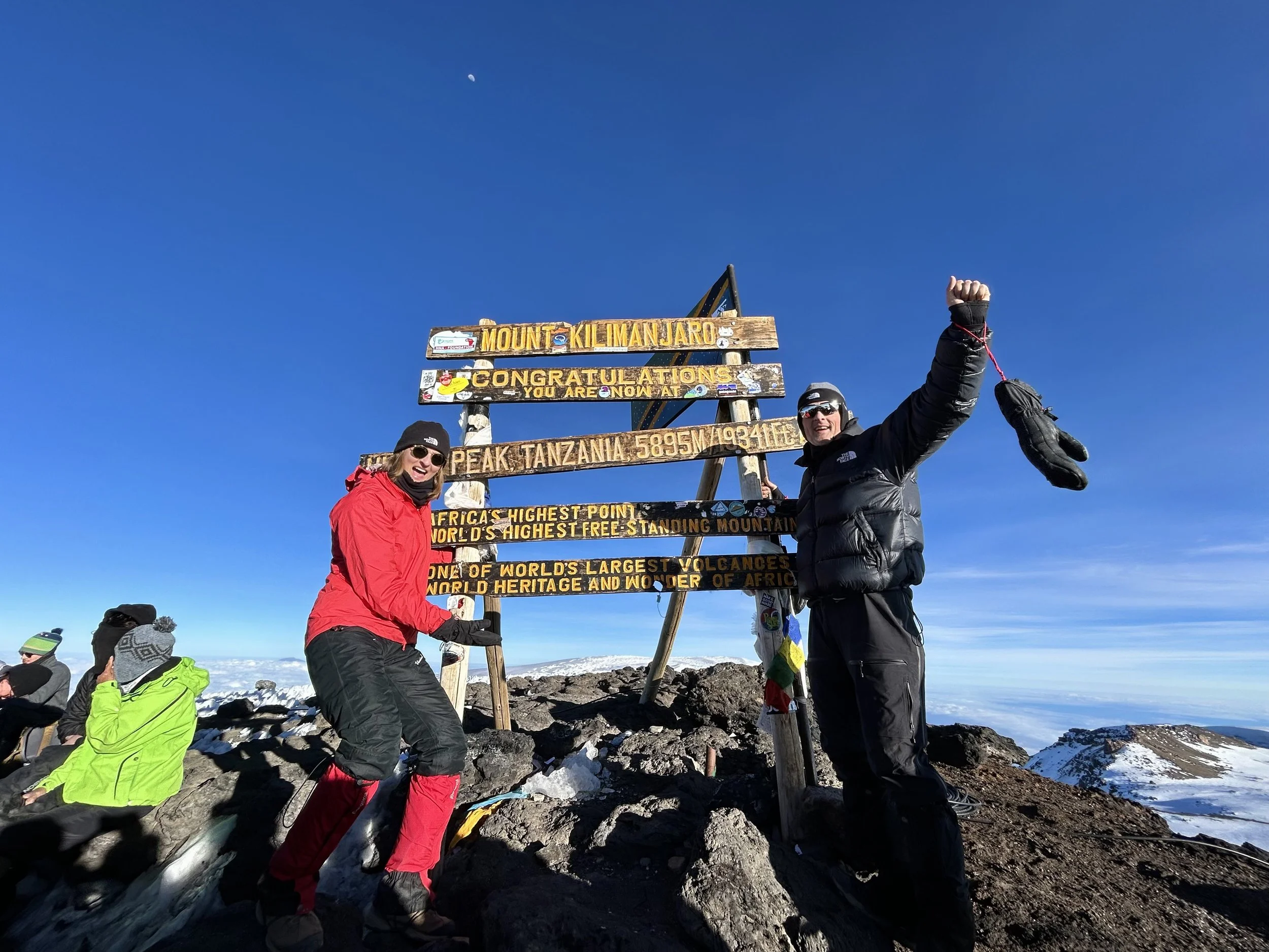 Two hikers standing next to a sign at the summit of Mount Kilimanjaro in Tanzania. One is in a red jacket and the other in a black jacket, both smiling and celebrating their achievement. The sign reads "Mount Kilimanjaro, Congratulations, Peak Tanzan